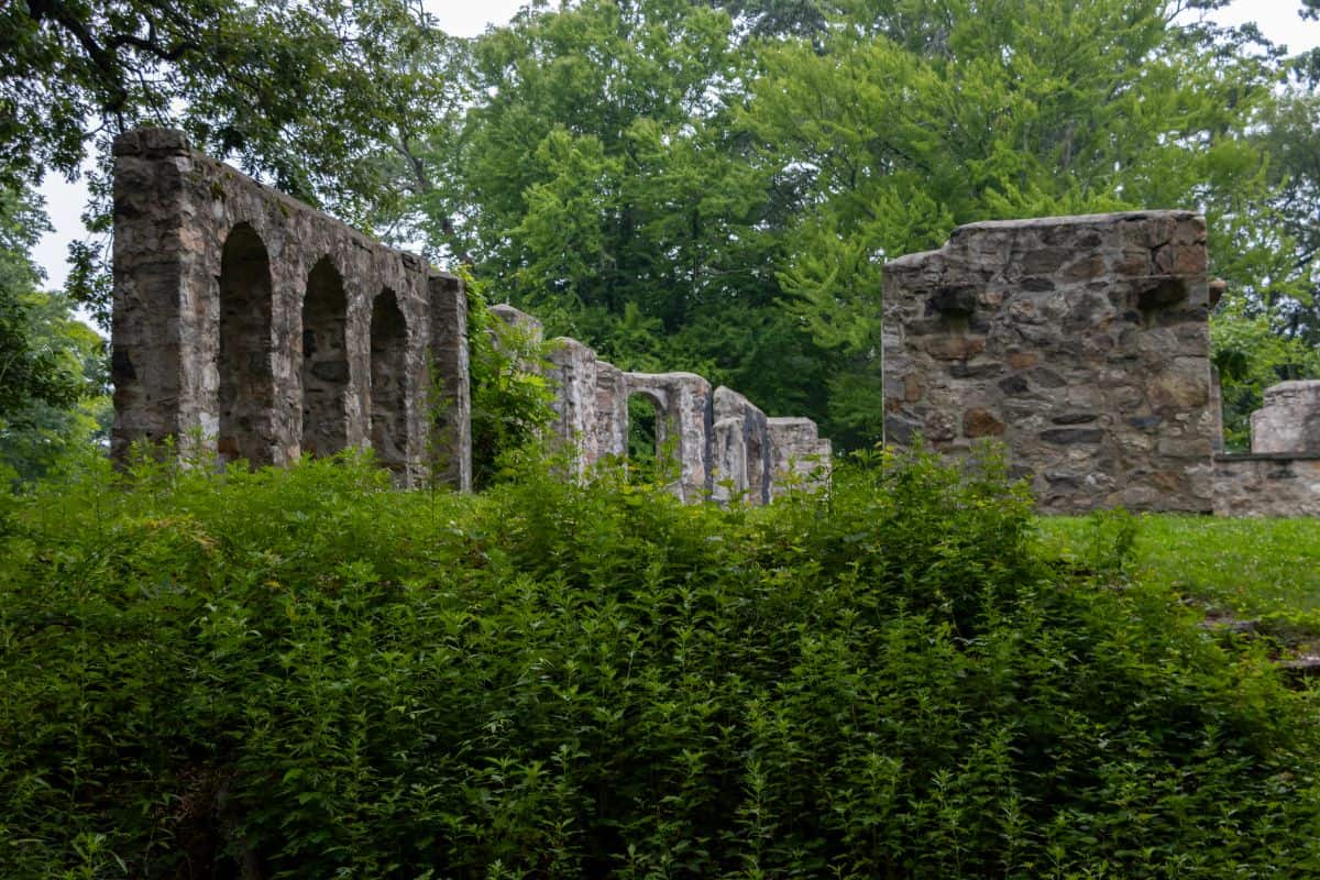 Stone ruins with arched openings stand amid green foliage and trees, suggesting remains of an old building in a forested area.