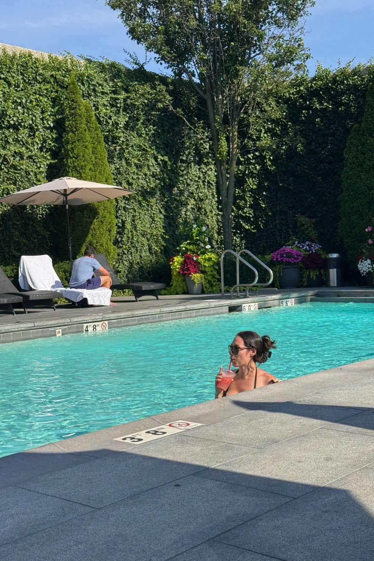 A woman in sunglasses holds a drink while standing in a pool; lounge chairs, an umbrella, and greenery are in the background.