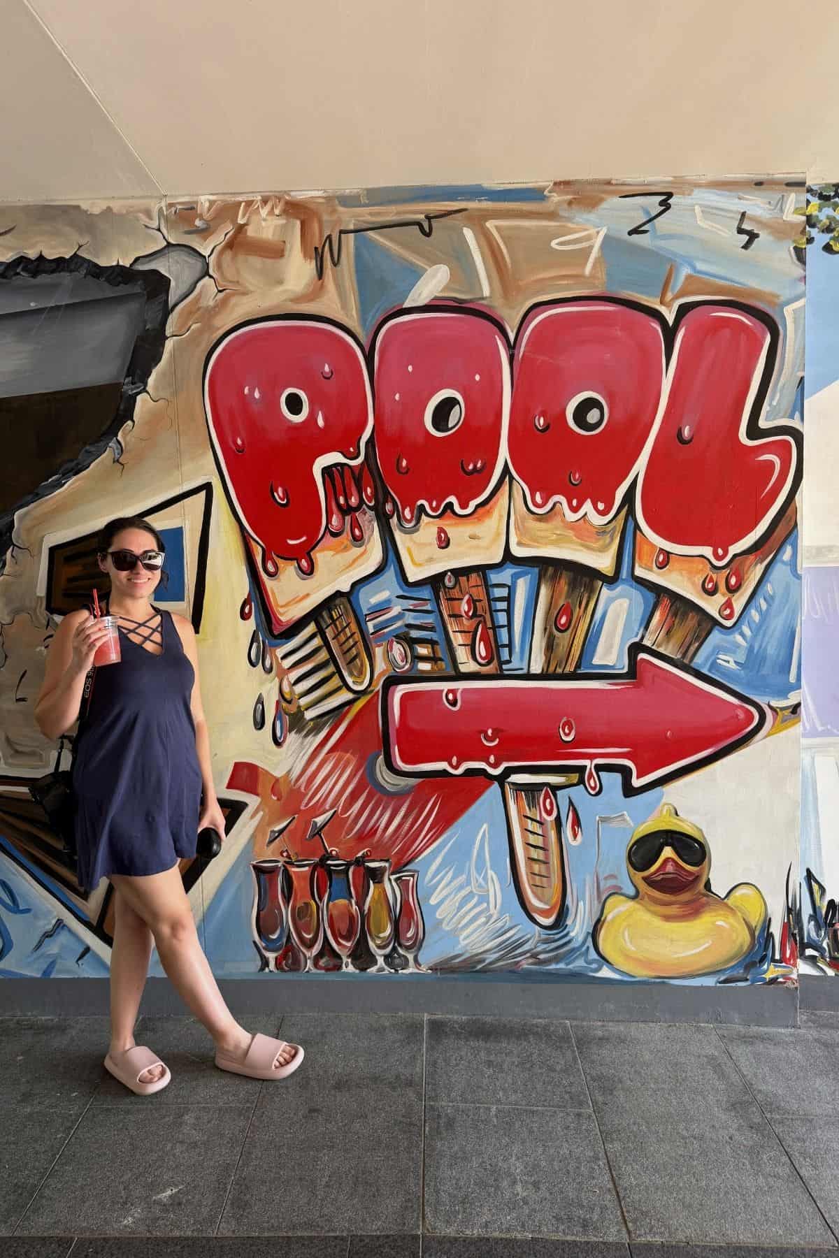 Woman in sunglasses holding a drink stands in front of a colorful mural with "POOL" written as popsicles and a red arrow pointing right. A rubber duck and drinks are painted at the bottom.