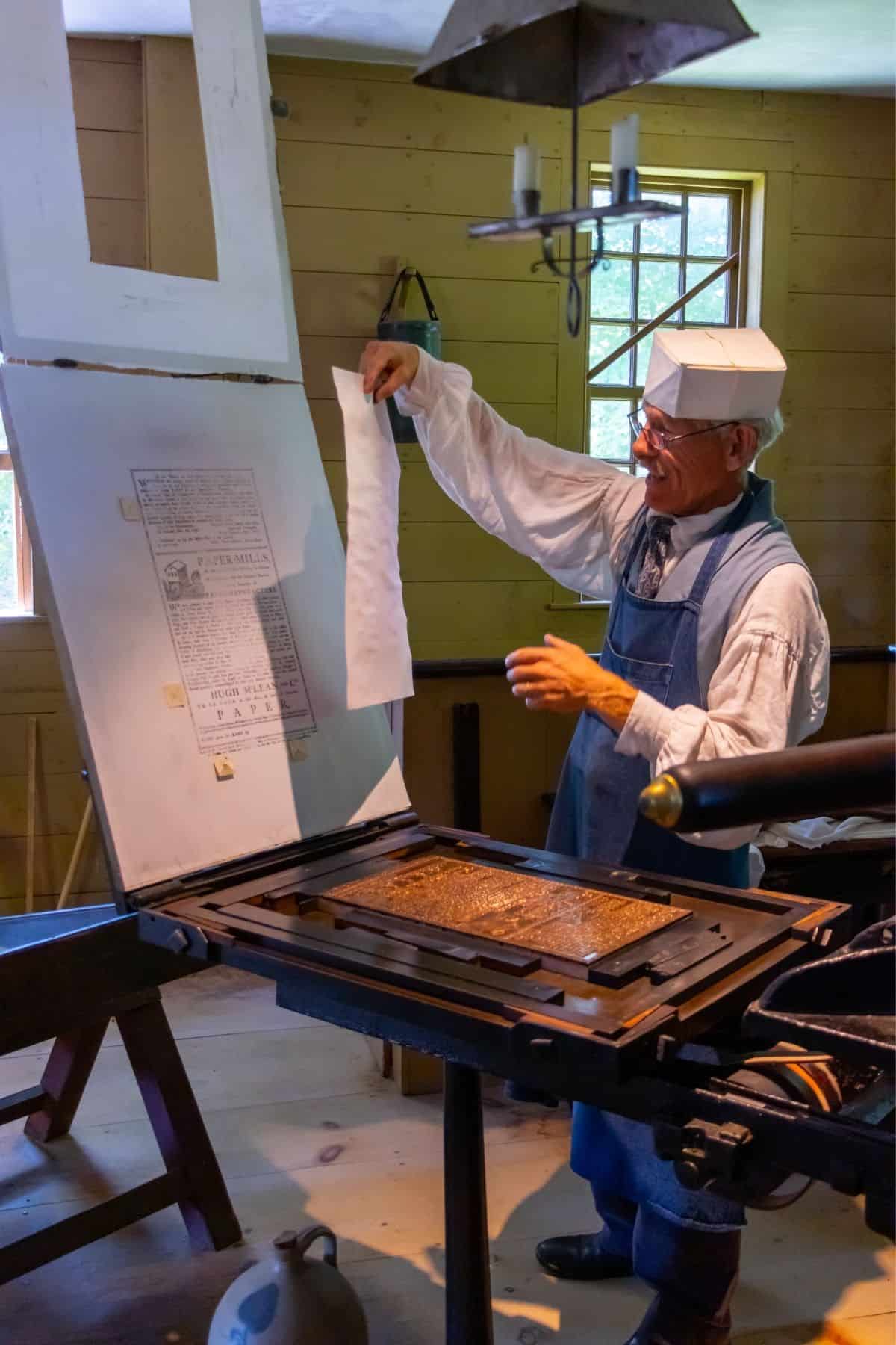 A man in historical clothing demonstrates an old-fashioned printing press inside a wooden room, holding a freshly printed sheet of paper.