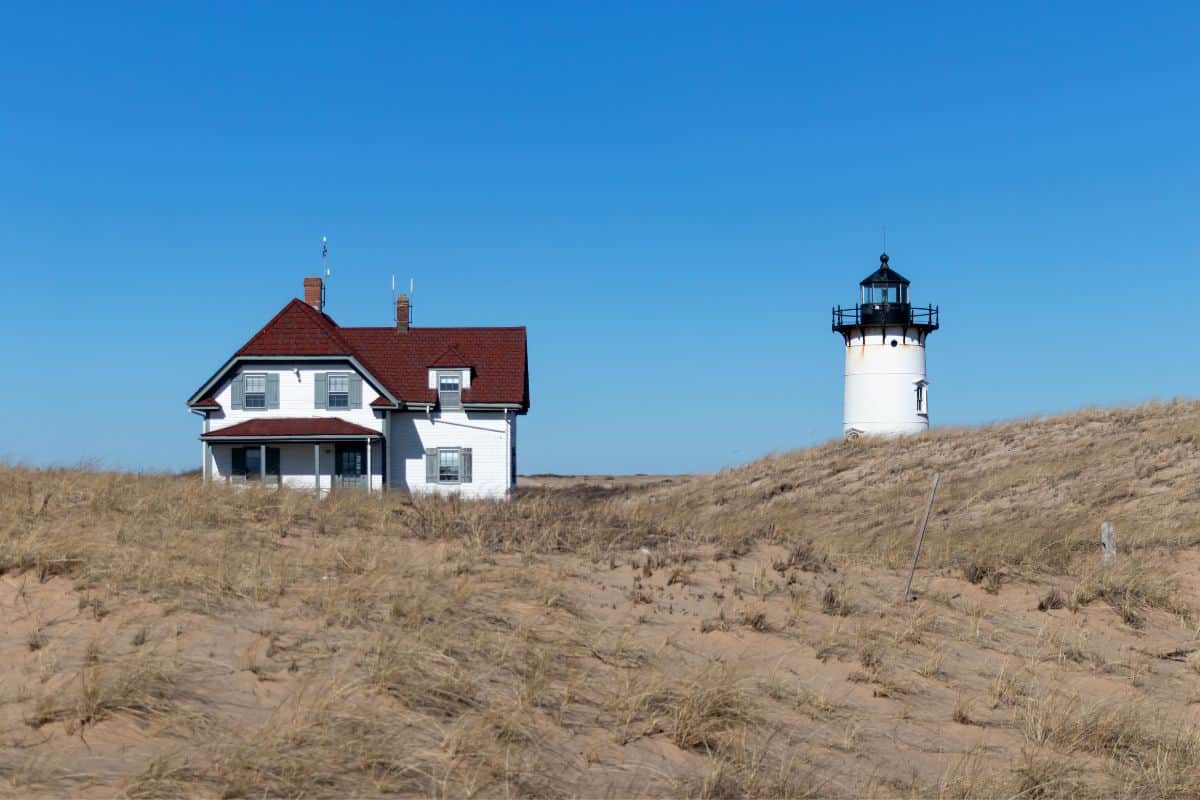 A white house with a red roof stands next to a white lighthouse on a sandy grassy dune under a clear blue sky.