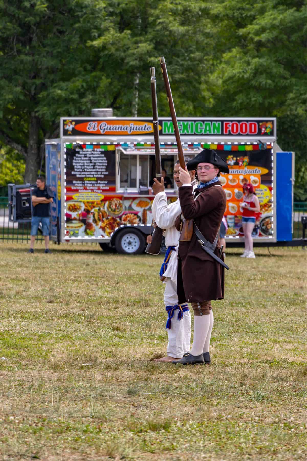 Two people in historical clothing stand with rifles on a grassy field, with a colorful Mexican food truck and several people in the background.