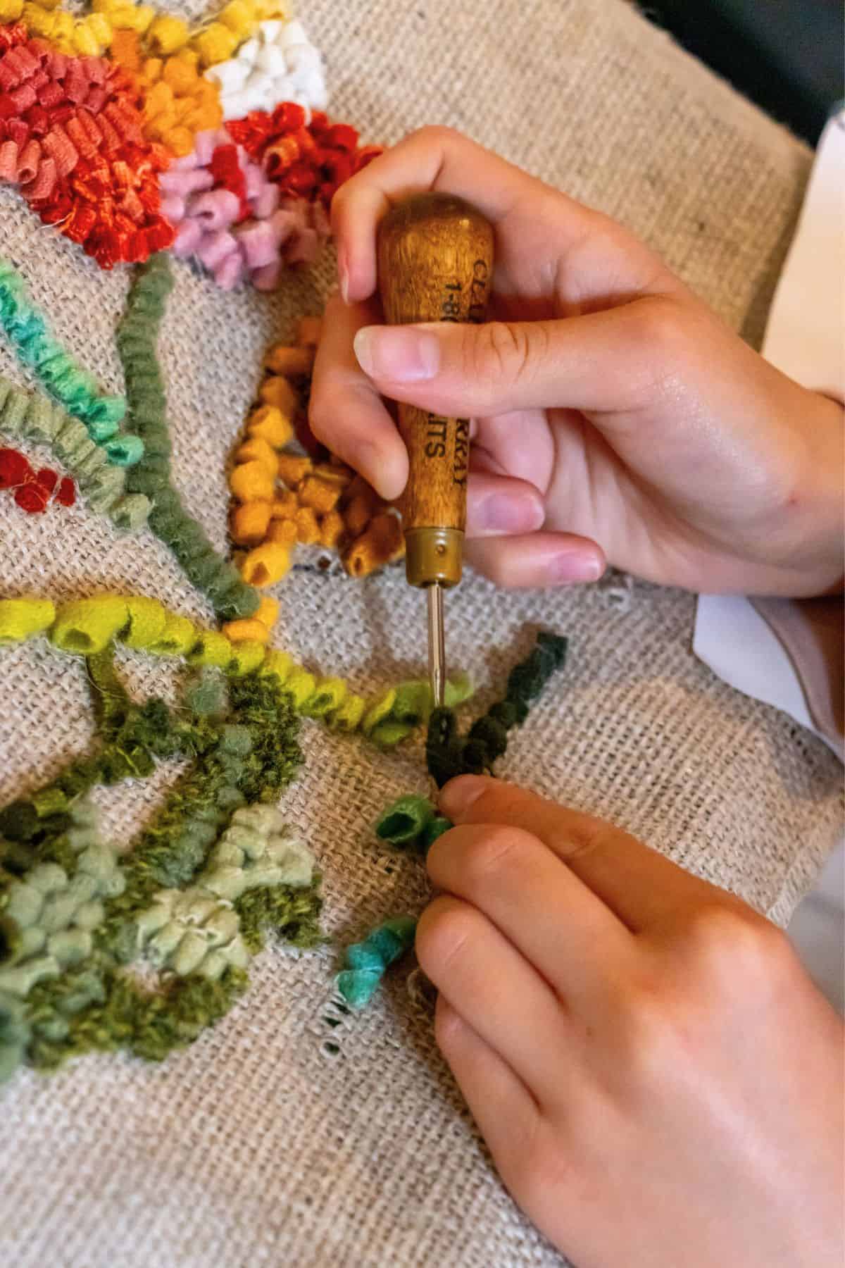 A person uses a wooden-handled punch needle to create a floral design with green and red yarn on burlap fabric.