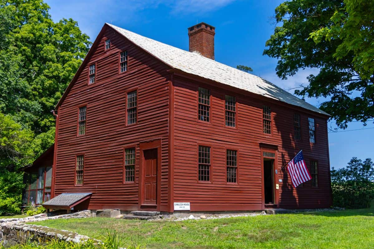 A large red wooden colonial-style house with many windows, a central brick chimney, and an American flag by the entrance, surrounded by green grass and trees under a blue sky.