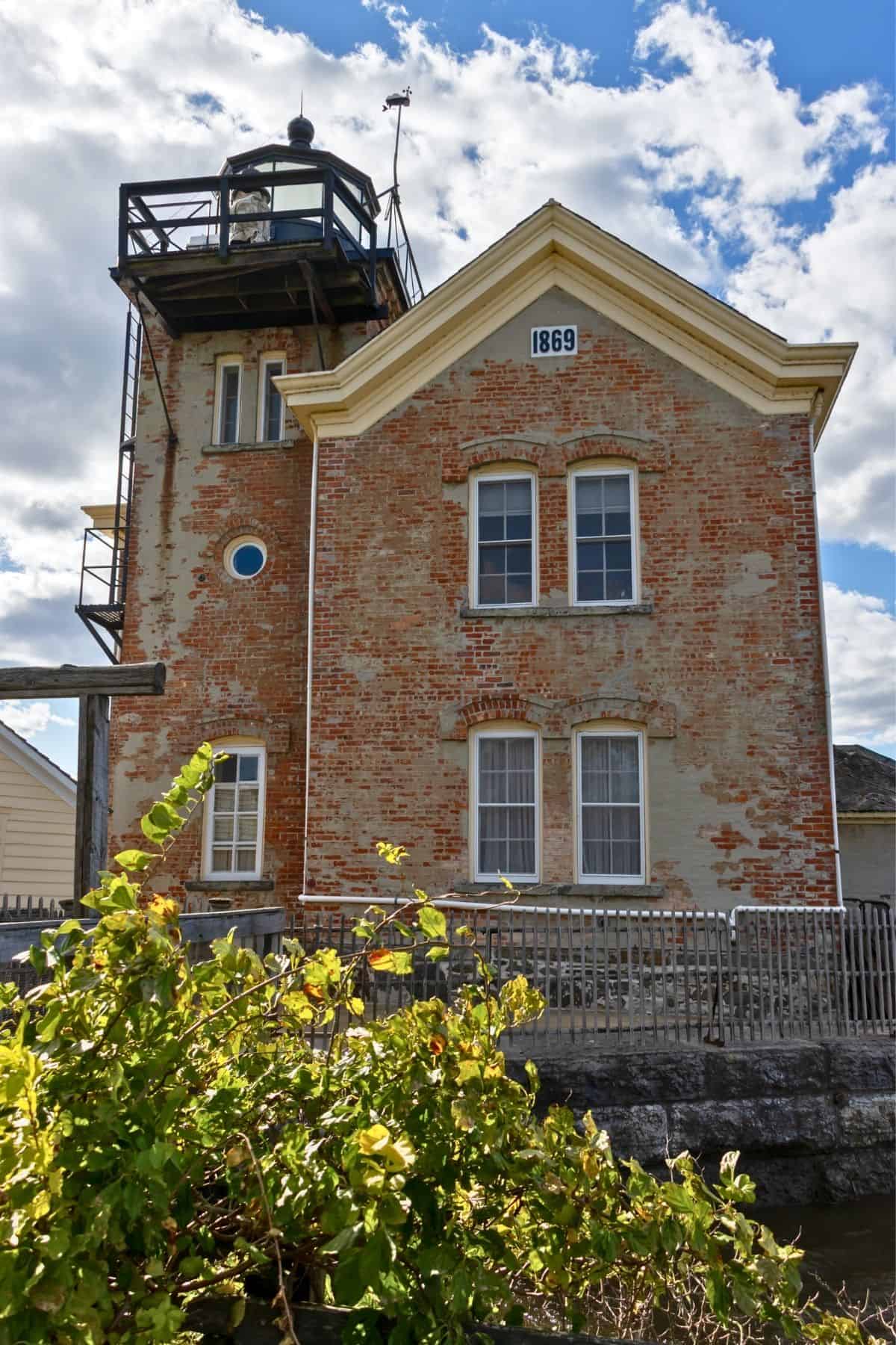 A brick lighthouse with "1869" on the façade, two arched windows, a balcony near the top, and greenery in the foreground under a partly cloudy sky.
