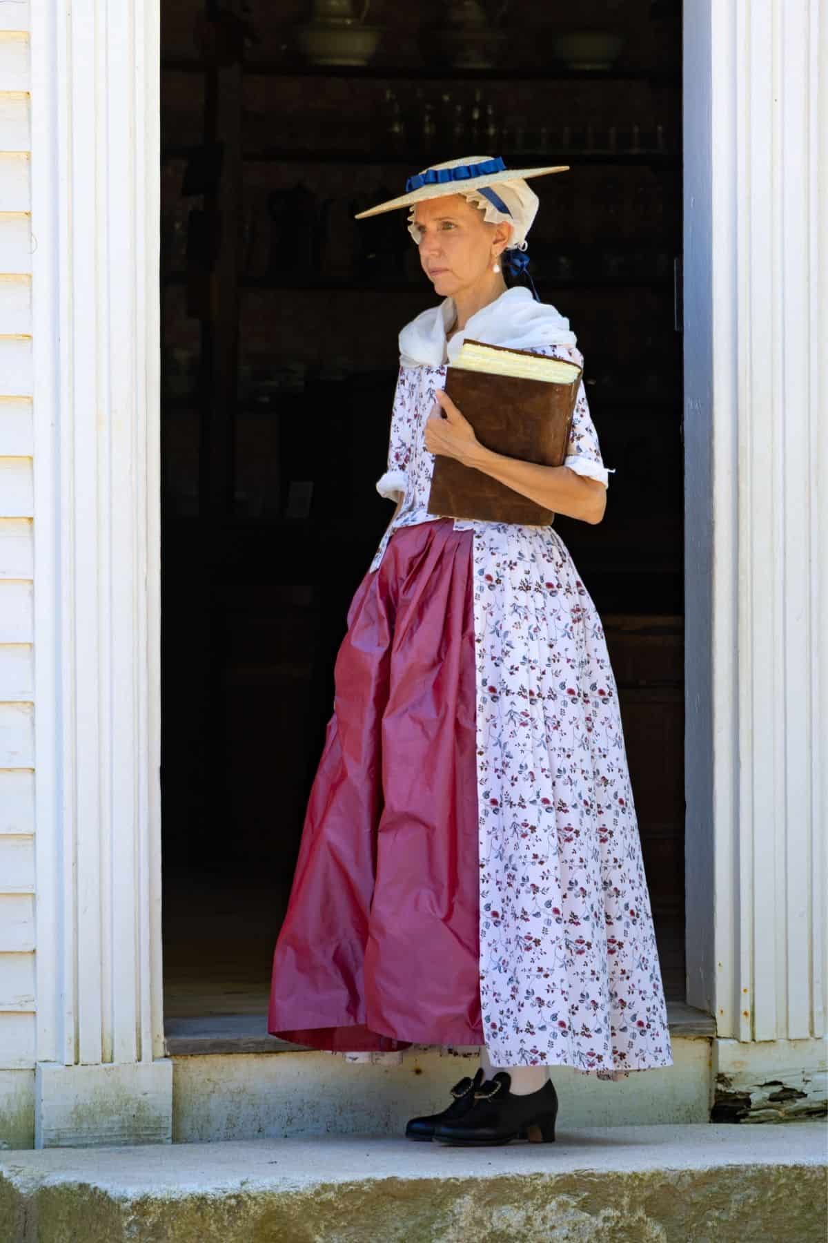 A woman in a historical costume stands in a doorway, holding a large book. She wears a long floral dress, a red apron, a headscarf, and a straw hat with a blue ribbon.