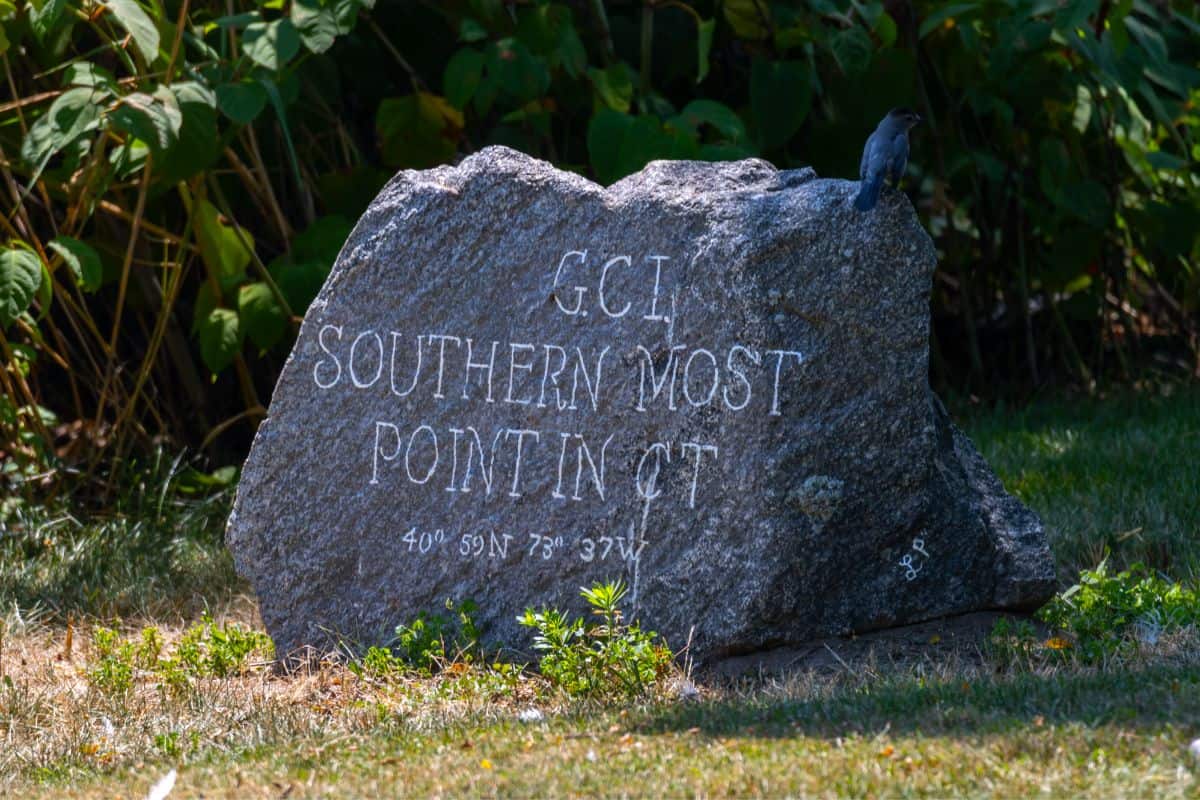 Large rock engraved with "GCI Southernmost Point in CT" and geographic coordinates, situated outdoors with green foliage in the background.