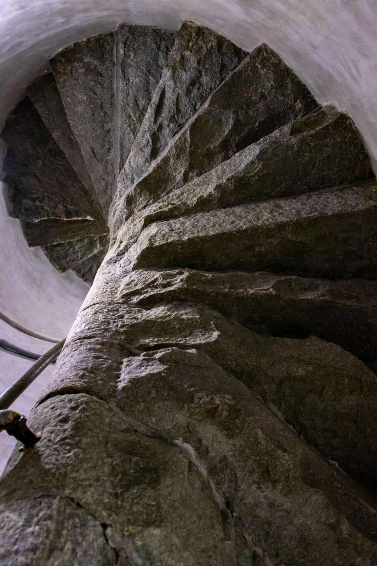 A stone spiral staircase viewed from below, showing rough textured steps winding upward within a narrow, cylindrical space.
