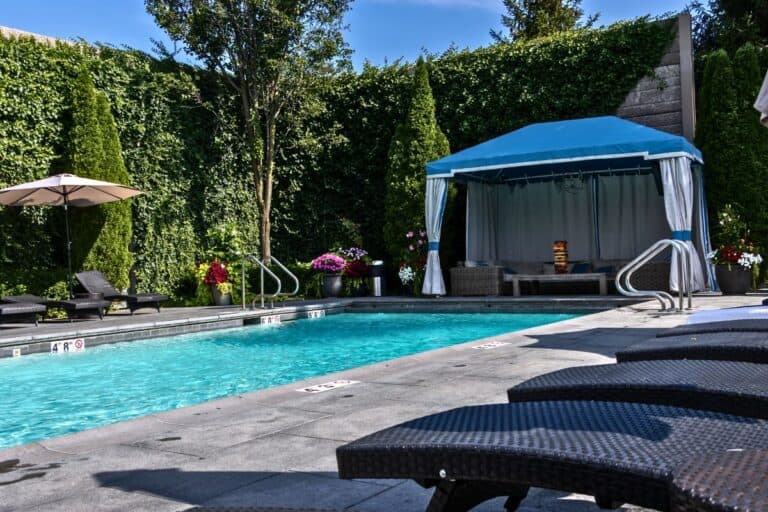 Rectangular outdoor swimming pool with lounge chairs, a blue canopy cabana, and tall green hedges. Sunlight creates a relaxed atmosphere.
