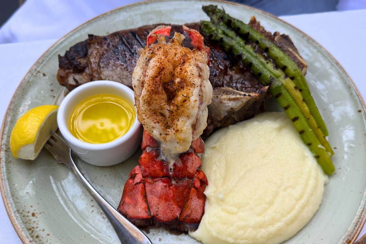 A plate with grilled steak, lobster tail, mashed potatoes, grilled asparagus, a lemon wedge, and a small dish of melted butter.