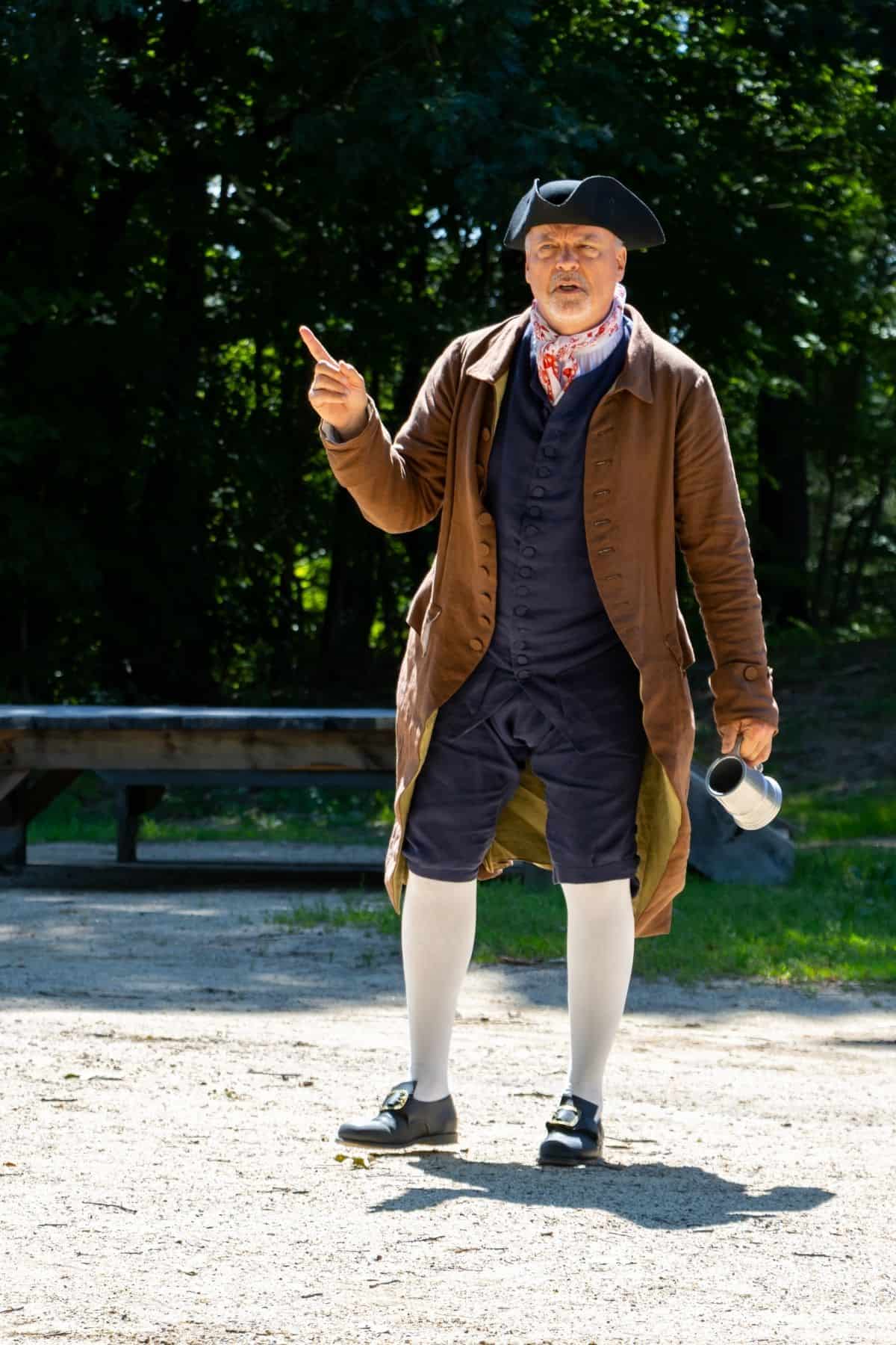 A man in colonial-era clothing stands outdoors, holding a metal mug in one hand and gesturing with the other.