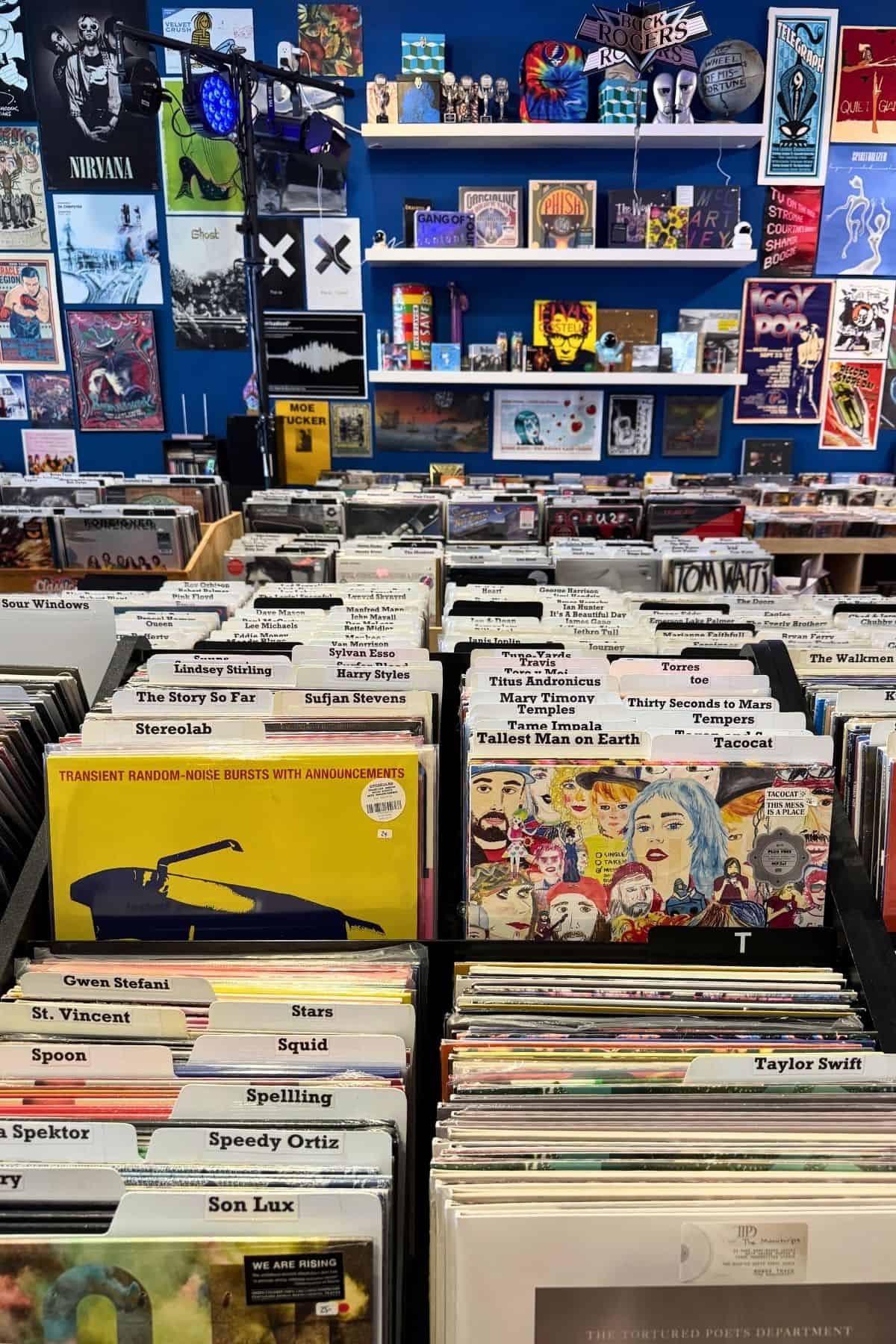Interior of a record store with vinyl records in bins and music-themed posters and album covers displayed on a blue wall in the background.