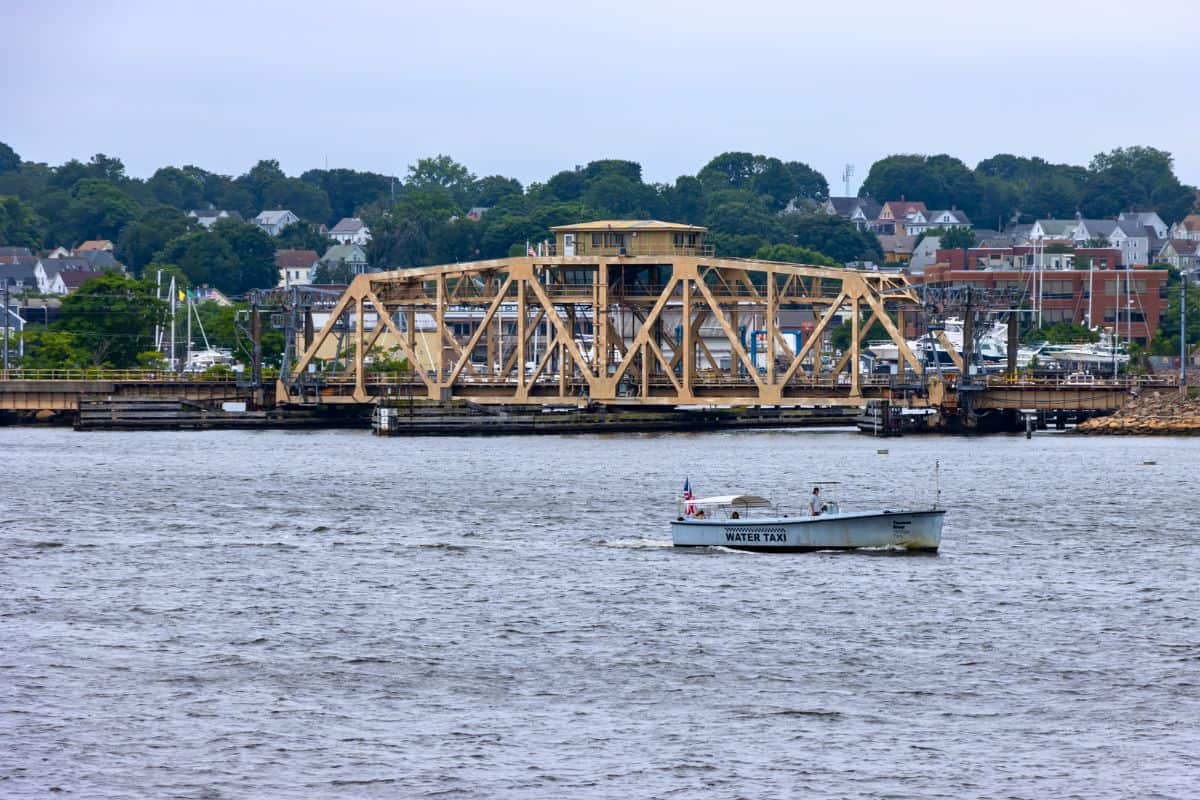 A small boat labeled "Water Taxi" moves across a river in front of a yellow drawbridge, with buildings and trees visible in the background.