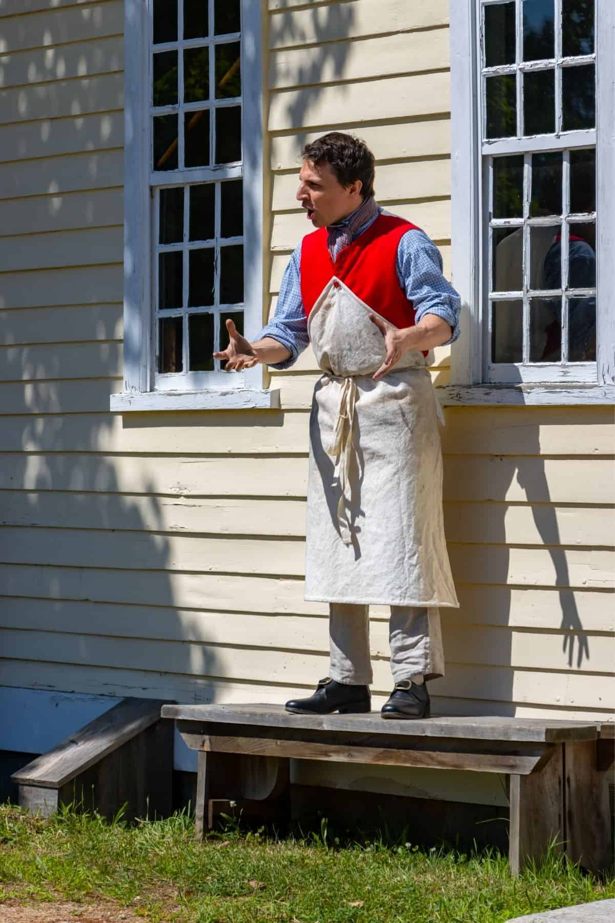 A person wearing a red vest and white apron stands on a bench outside a yellow building, gesturing with both hands.