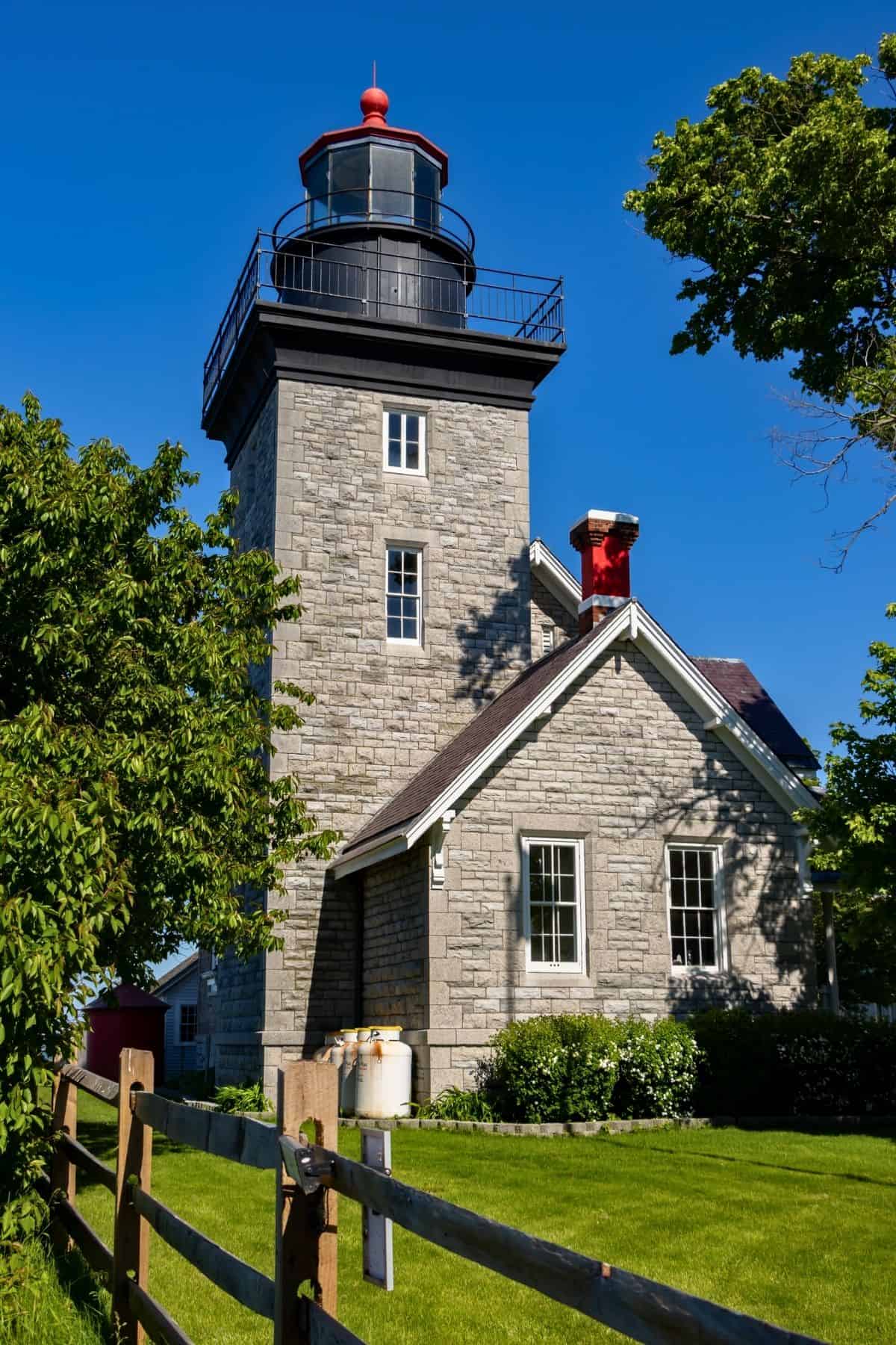 Stone lighthouse with black lantern and red roof accents, surrounded by green trees and lawn, under a clear blue sky.