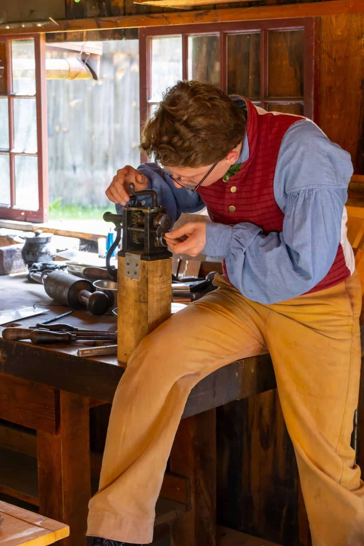 Person in historical clothing uses a small vise or hand tool on a wooden workbench in a rustic workshop with tools and open windows.