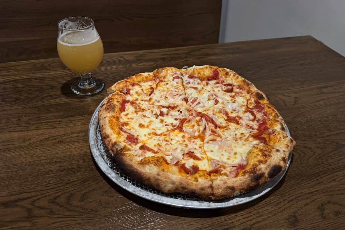 A whole pizza with cheese and onions sits on a metal tray on a wooden table next to a glass of pale beer.