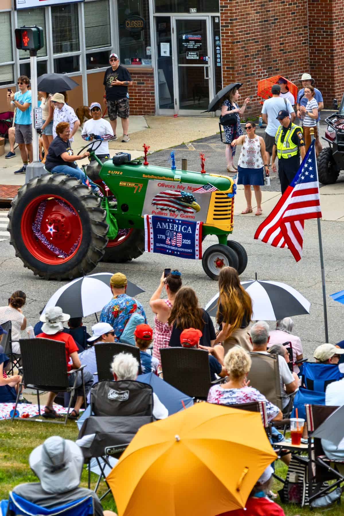 A green vintage tractor with American flags drives in a parade; people sit and stand along the street, many holding umbrellas.