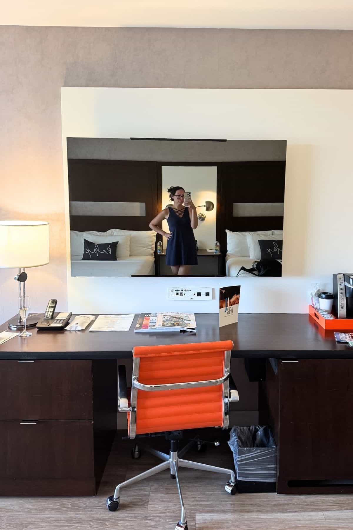 A person stands in front of a mirror taking a photo in a hotel room, with a desk, orange chair, lamp, and coffee station visible in the foreground.