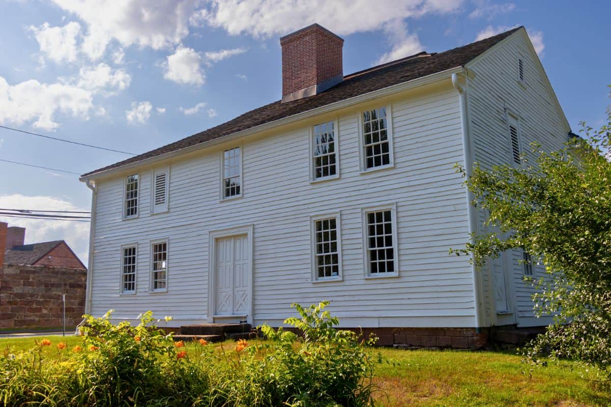 A white, two-story historic house with many windows, a brick chimney, and a sloped roof stands on a grassy lawn under a partly cloudy sky.