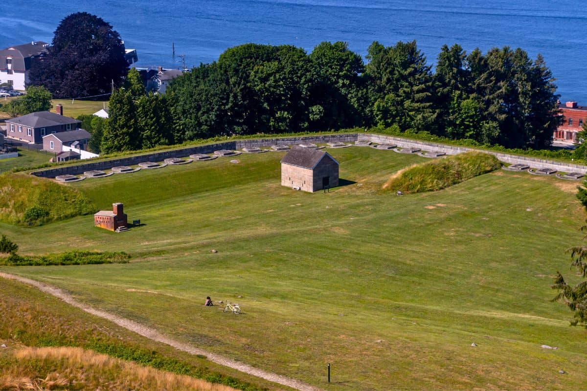 A rectangular grassy field with a stone wall, small central building, scattered ruins, trees, and water in the background.