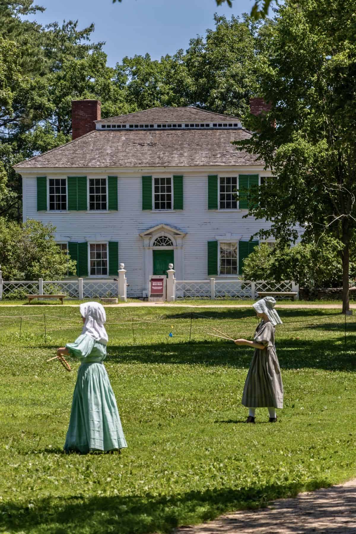Two people in historical clothing play croquet on a lawn in front of a large white house with green shutters and a fenced yard.