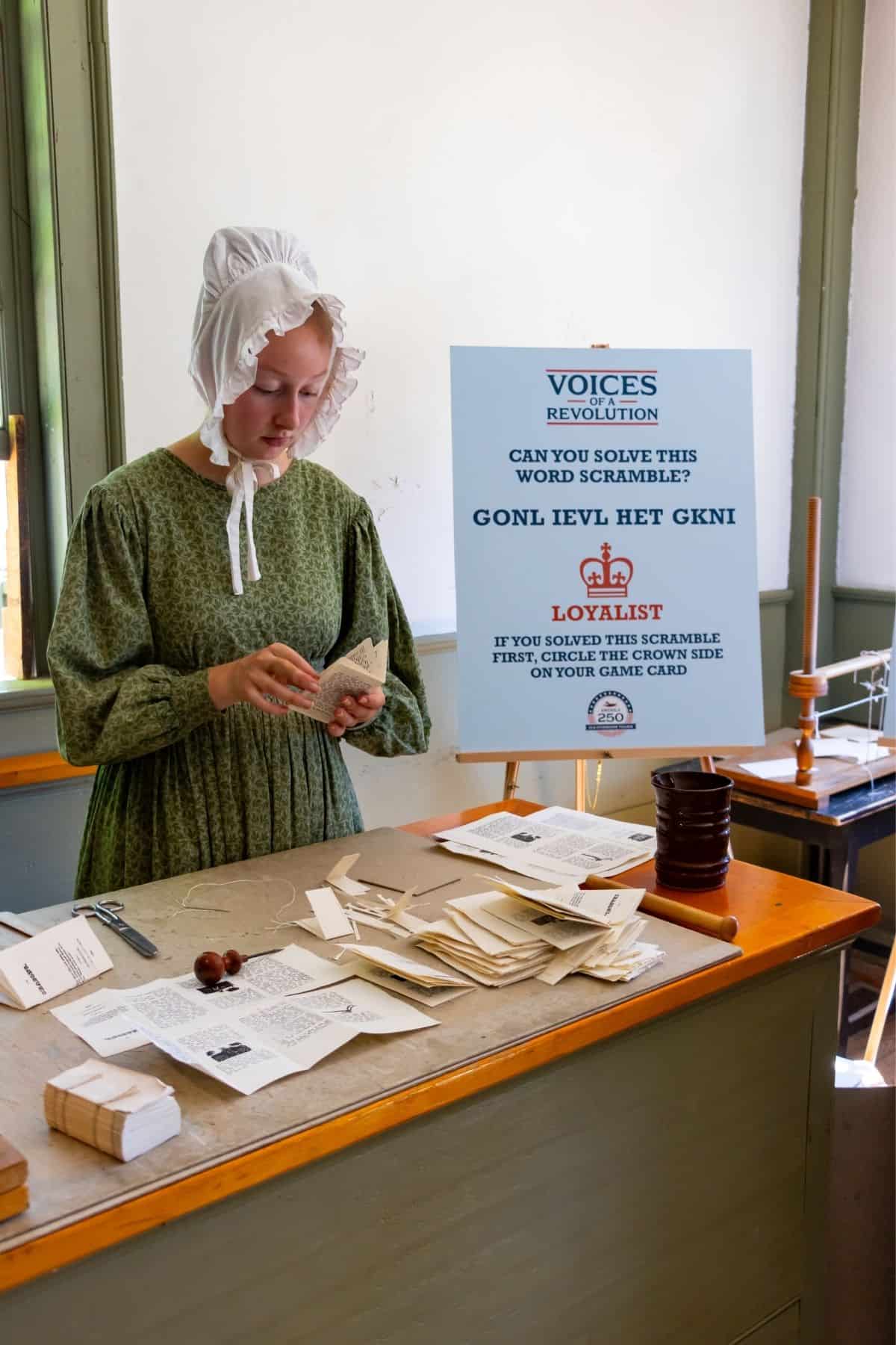 A woman in historical clothing stands at a table sorting papers. Behind her is a sign with a word scramble and instructions for a game related to the American Revolution.