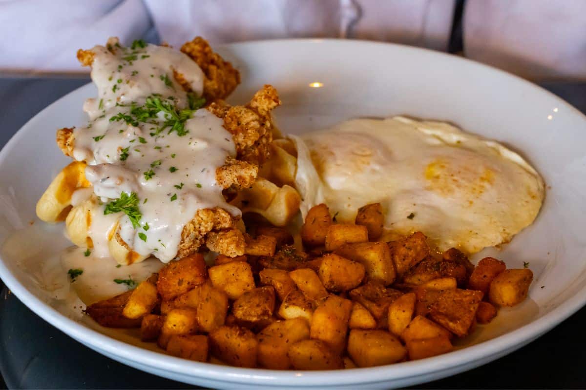 A plate of chicken and waffles with gravy, two fried eggs, and seasoned diced potatoes.
