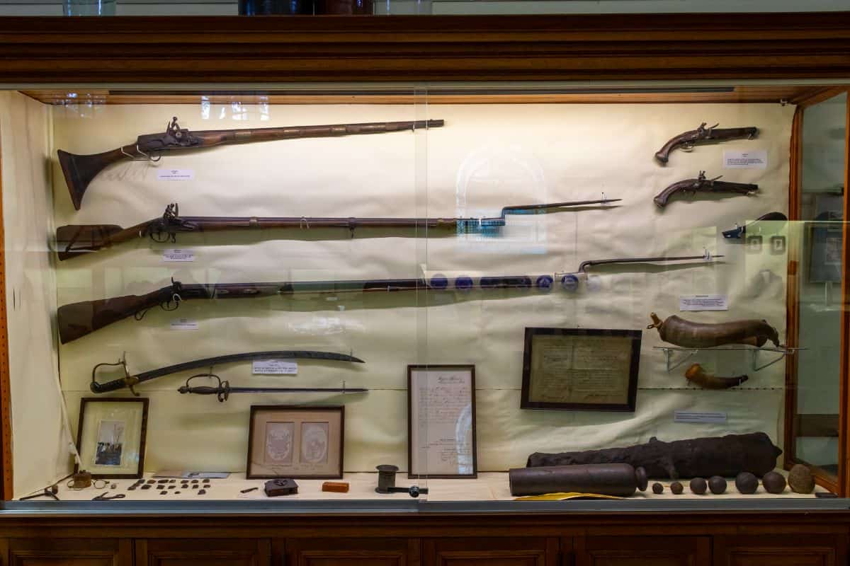 A glass display case exhibiting antique rifles, pistols, bayonets, swords, framed documents, and various small artifacts in a museum setting.
