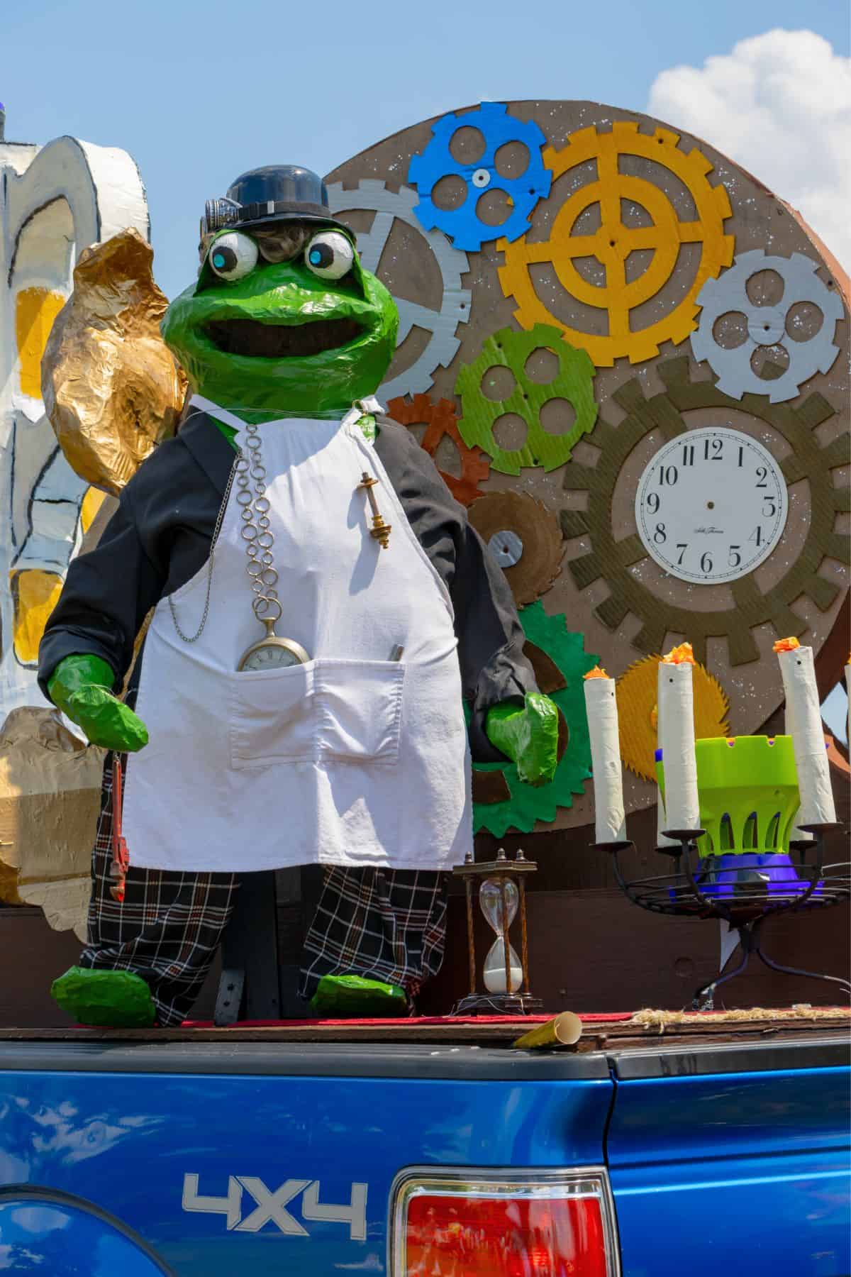A frog statue dressed as a chef with a clock, large gears, and candles stands on the back of a blue 4x4 truck during a parade or outdoor event.