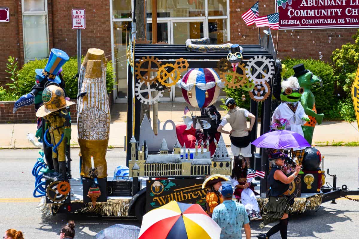 A parade float decorated with steampunk elements and costumed participants passes by a church building; people with umbrellas watch from the street.