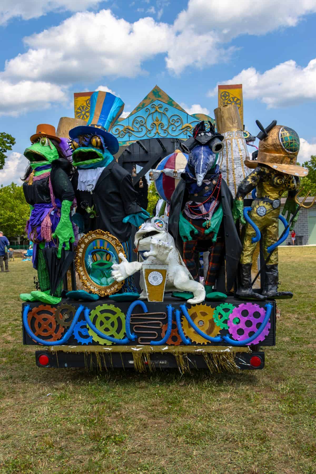 A parade float decorated with large, colorful frog sculptures in steampunk attire stands on a grassy field under a partly cloudy sky.