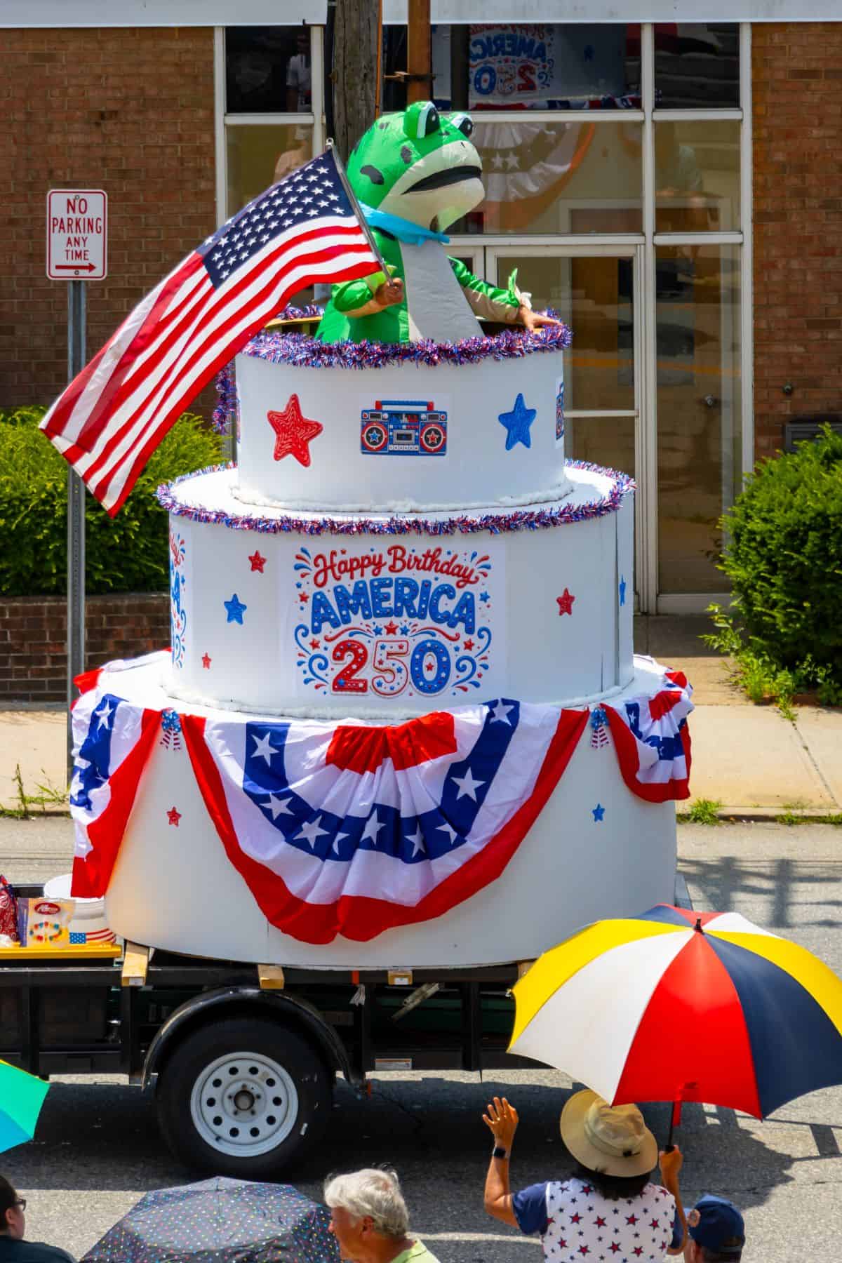 A parade float features a large birthday cake prop with patriotic decorations, an American flag, and a green dinosaur on top. People with colorful umbrellas watch from the street.