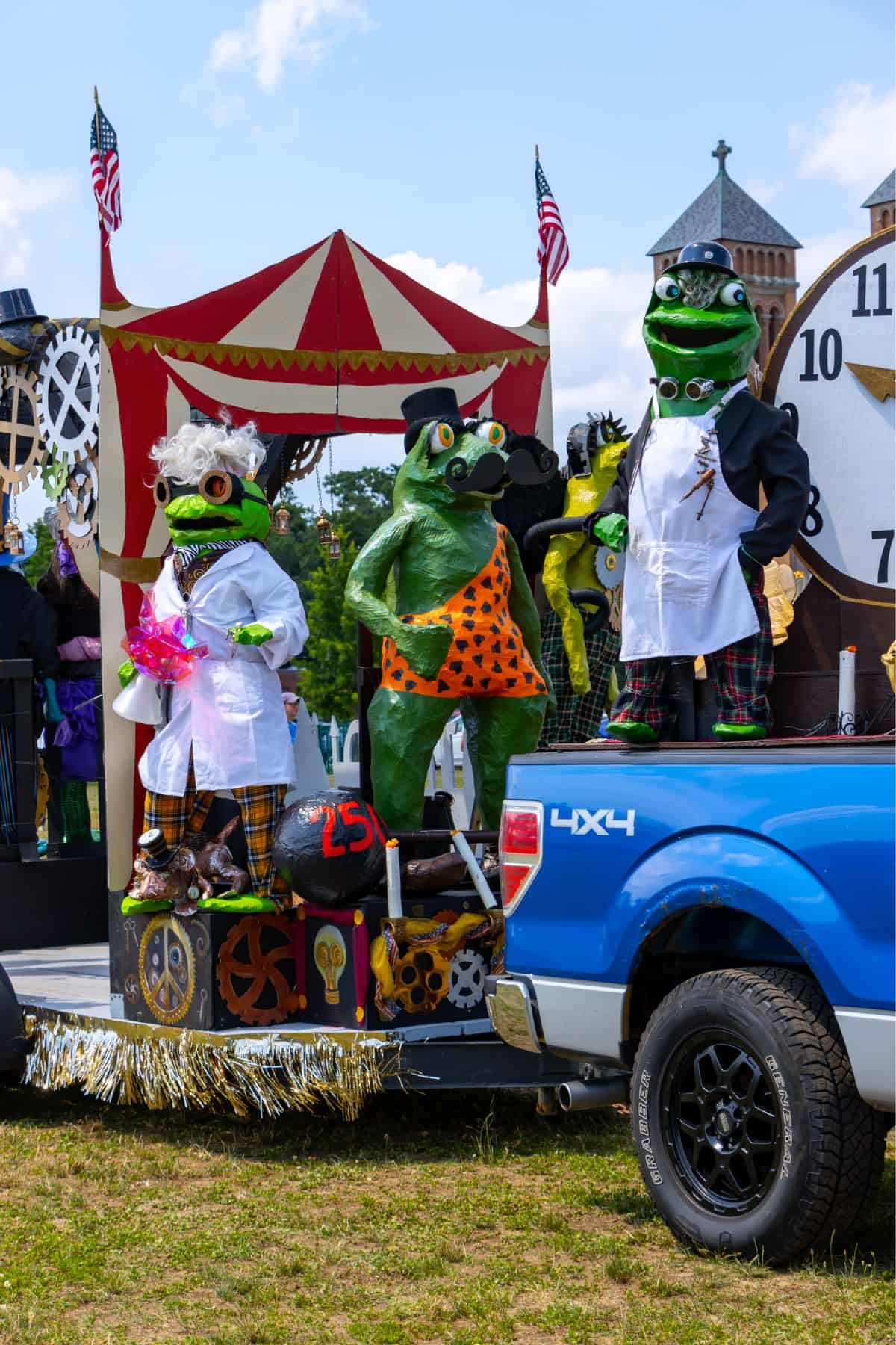 Three large frog statues in costumes stand on a parade float next to a blue pickup truck, with a carnival tent backdrop and steampunk decorations in the background.