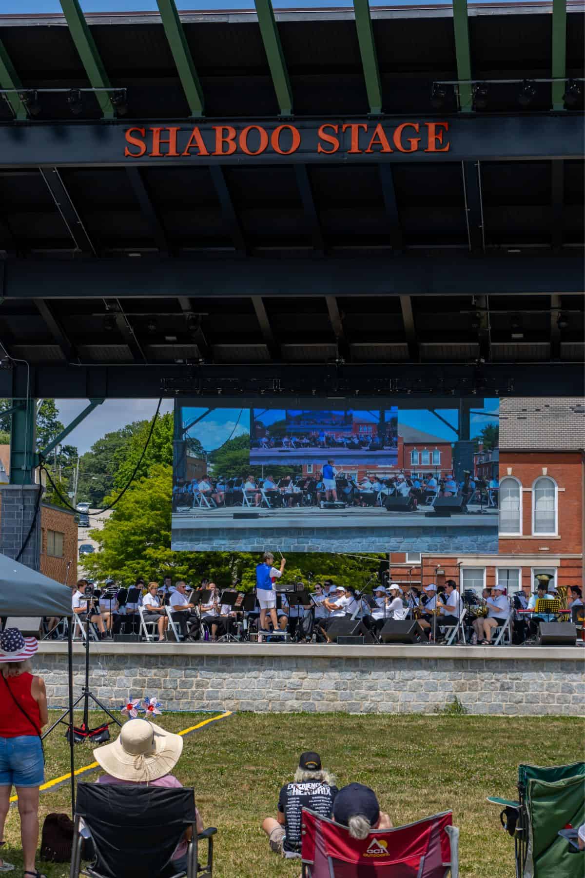 People sit on the grass watching a performance on Shaboo Stage, with a large screen displaying the musicians and conductor.