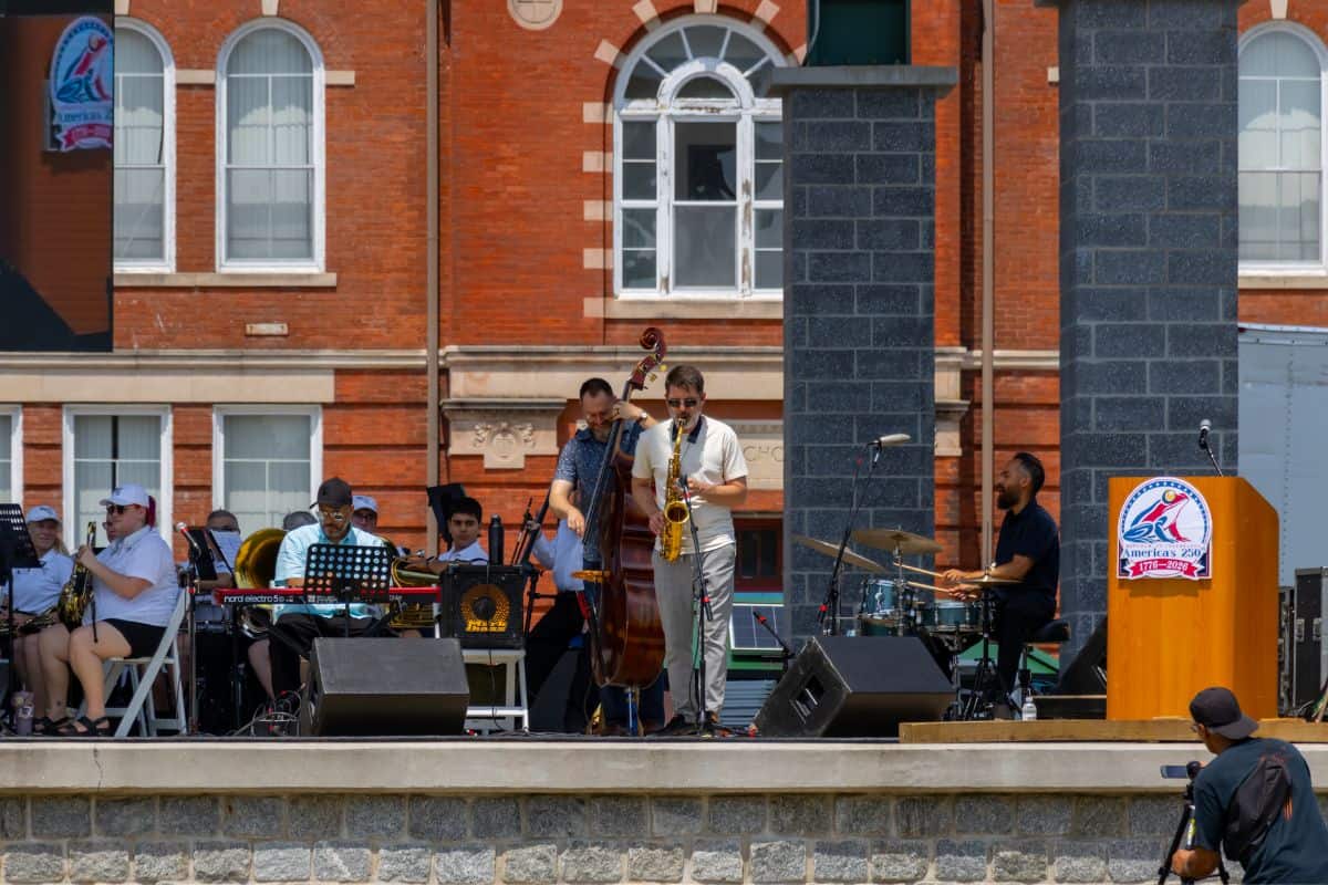 A jazz band performs on an outdoor stage with musicians playing keyboard, saxophone, upright bass, and drums in front of a brick building.