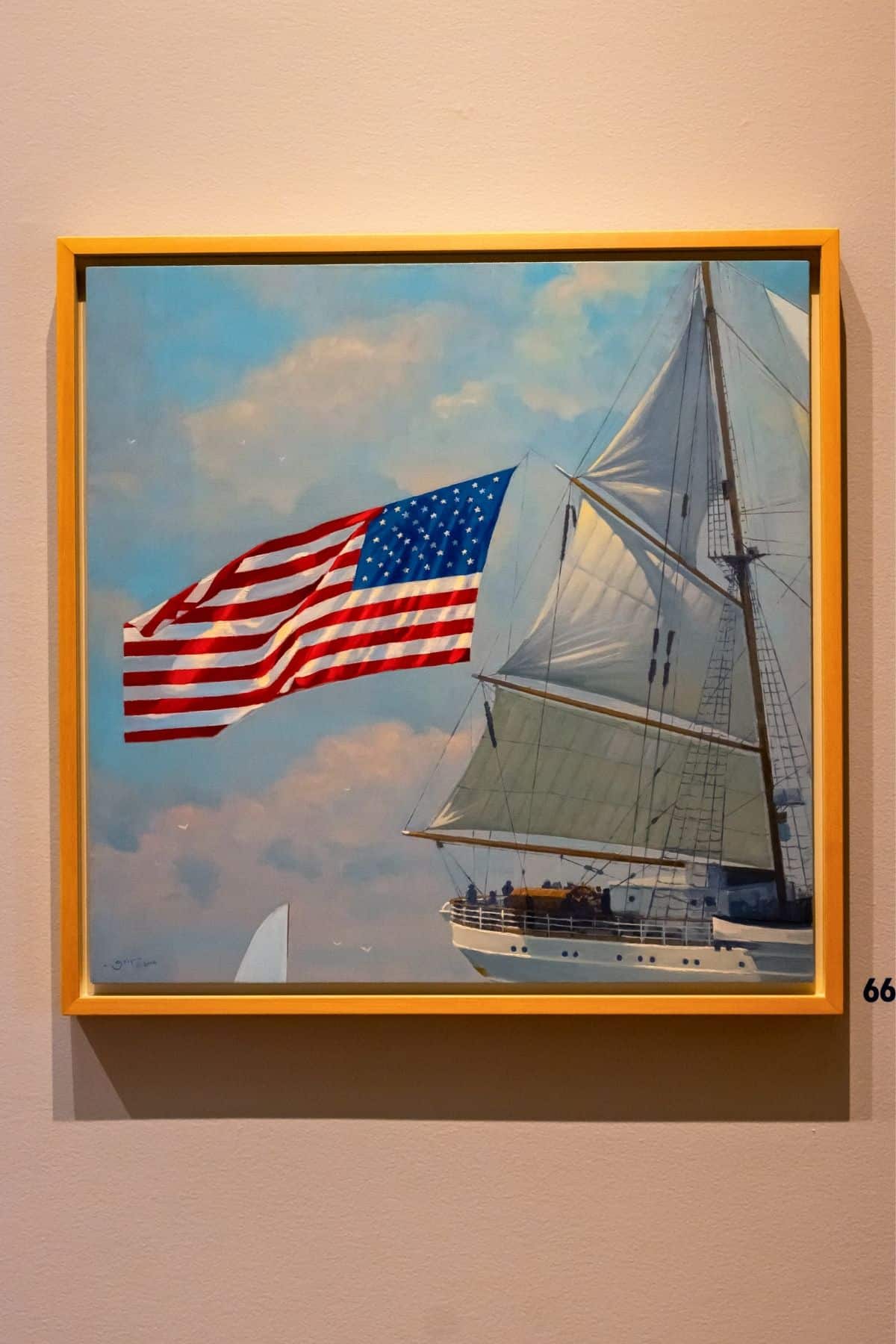 Framed painting of a tall sailing ship with a large American flag flying at the stern, mounted on a beige wall.