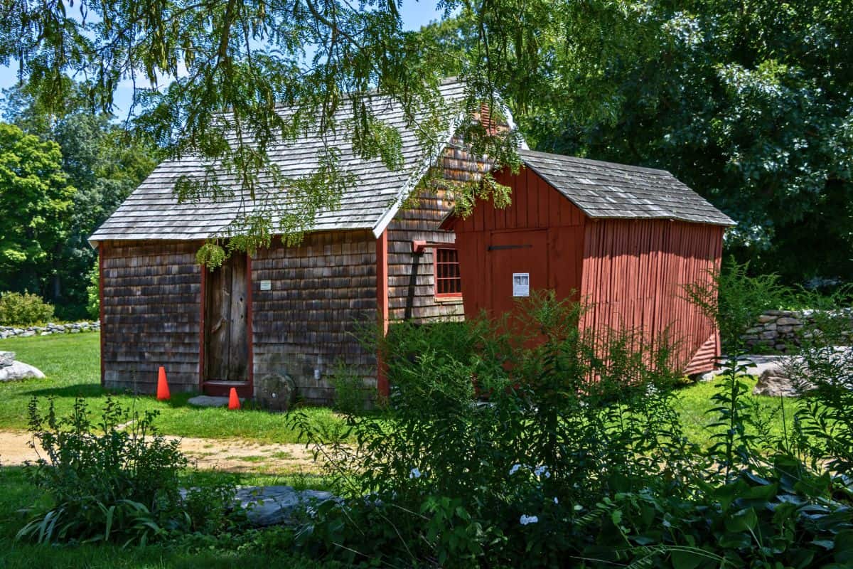 A small wooden building with shingle siding and a red extension stands on grass, partially shaded by trees. Two orange cones are near the door.
