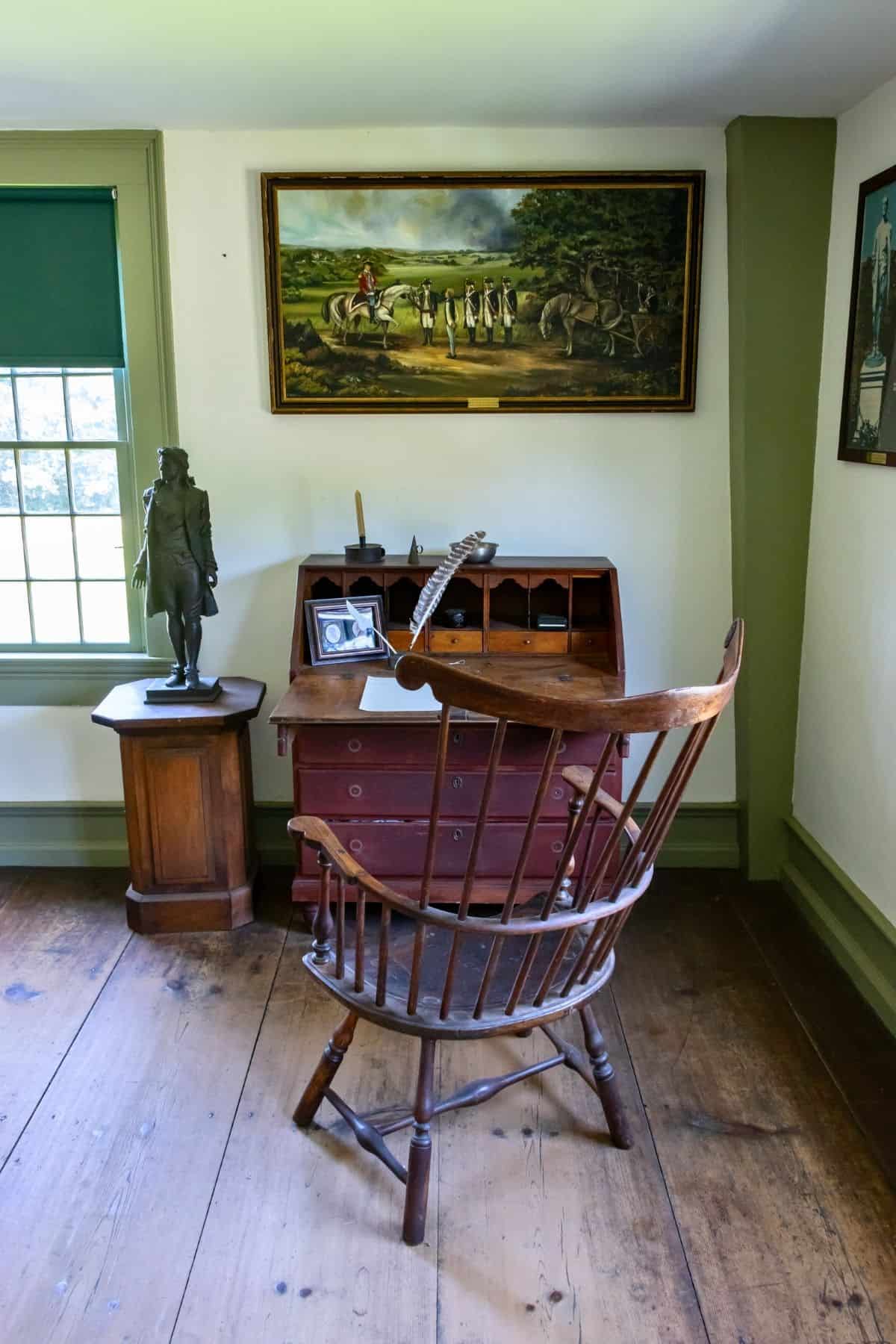 A wooden desk and chair sit by a window in a historic room, with a statue, framed photos, and a large painting of soldiers on the wall above.