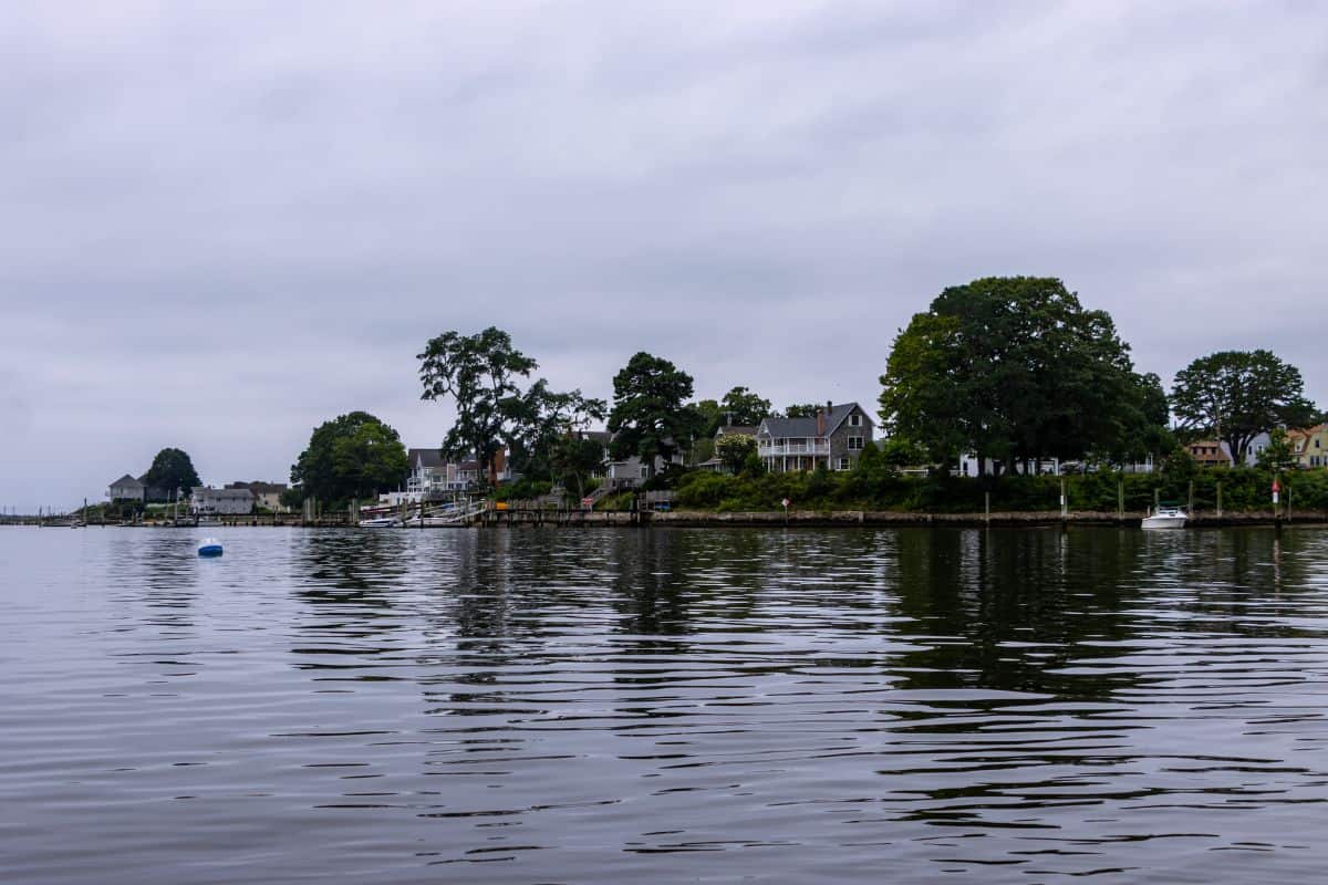 Houses and trees line a calm, cloudy shoreline, with their reflections visible in the still water. A blue buoy floats in the water near the shore.