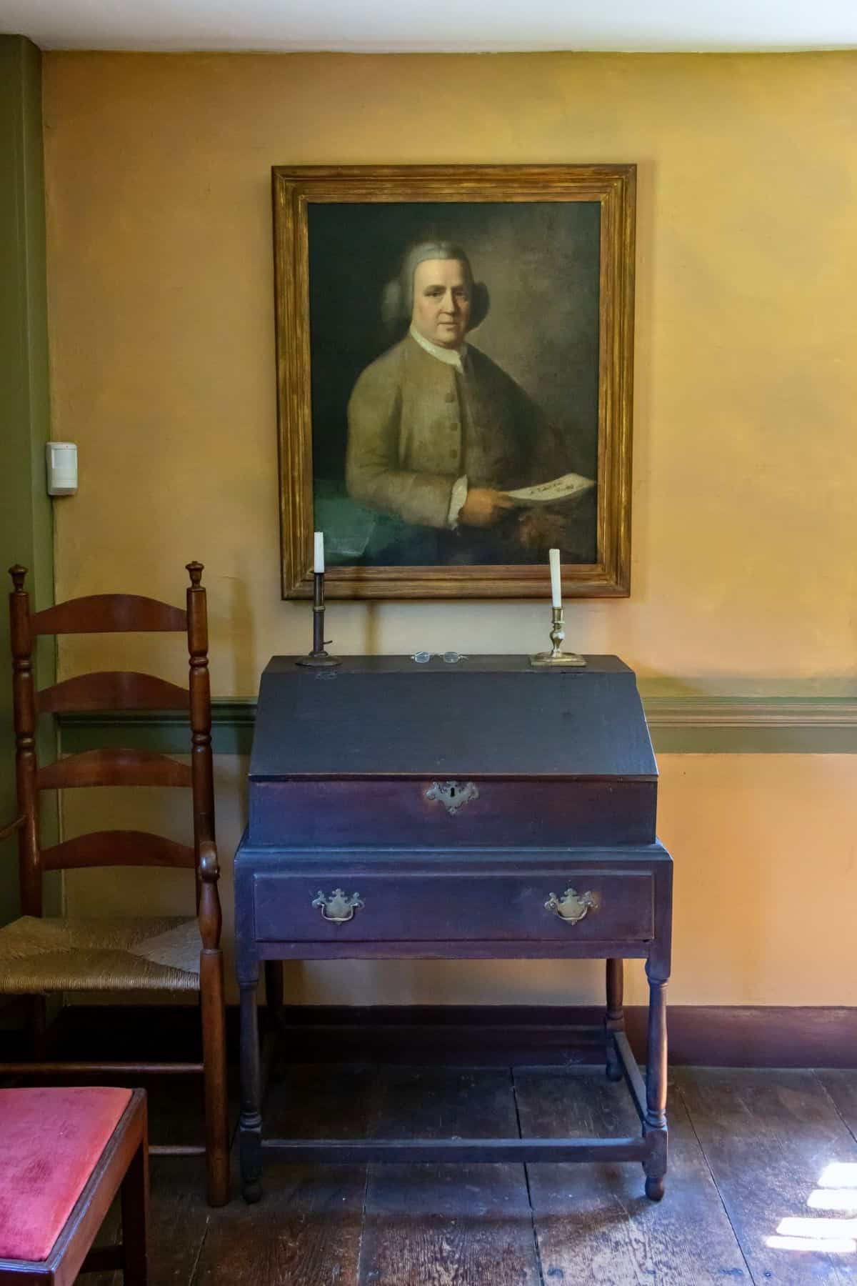 A wooden writing desk with two candlesticks sits beneath a framed portrait of a man in historical clothing on a tan wall.