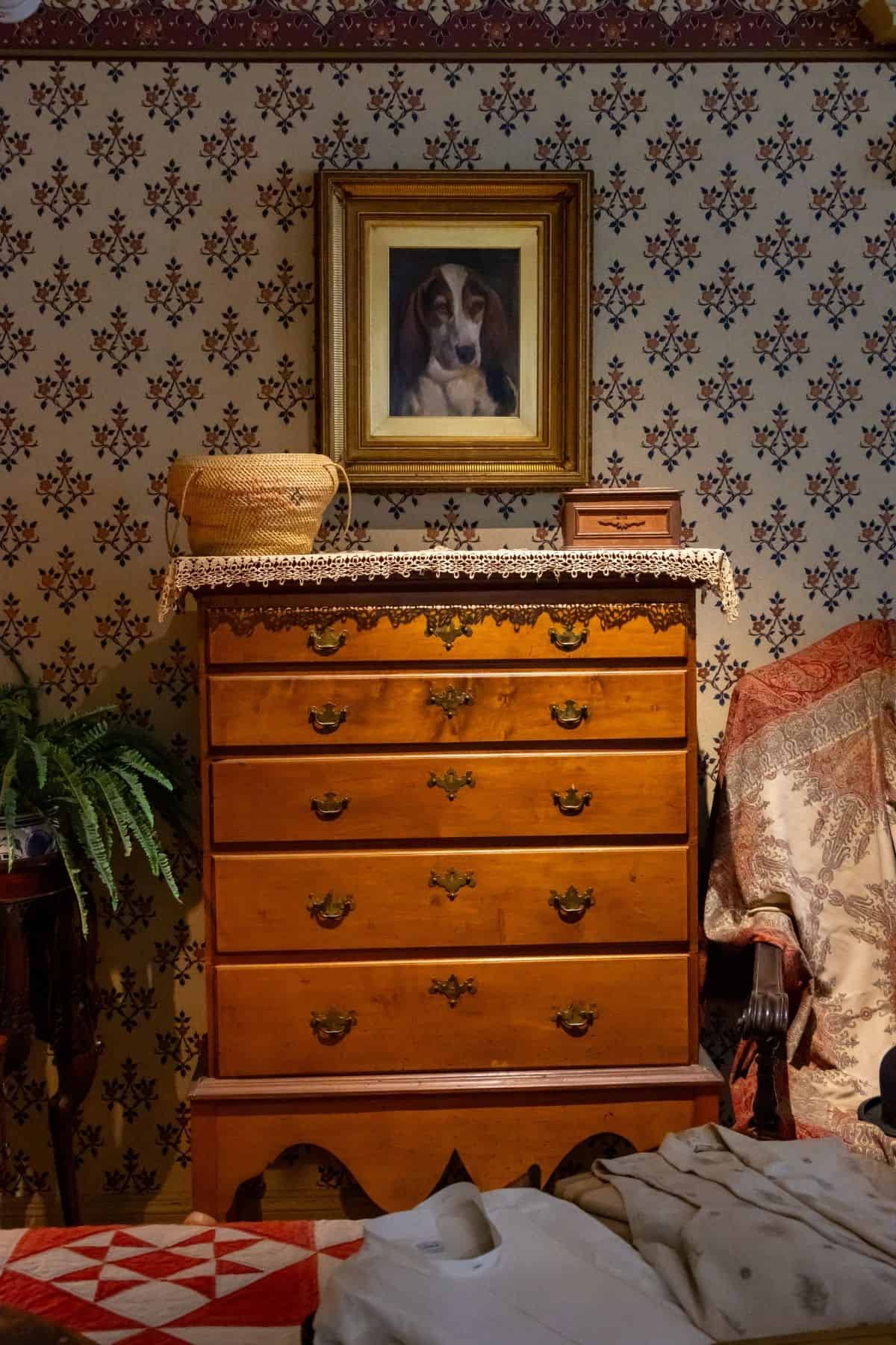 A wooden dresser with six drawers stands against floral wallpaper, topped with a lace doily, a basket, a box, and a framed dog portrait. A fern and fabric are nearby.