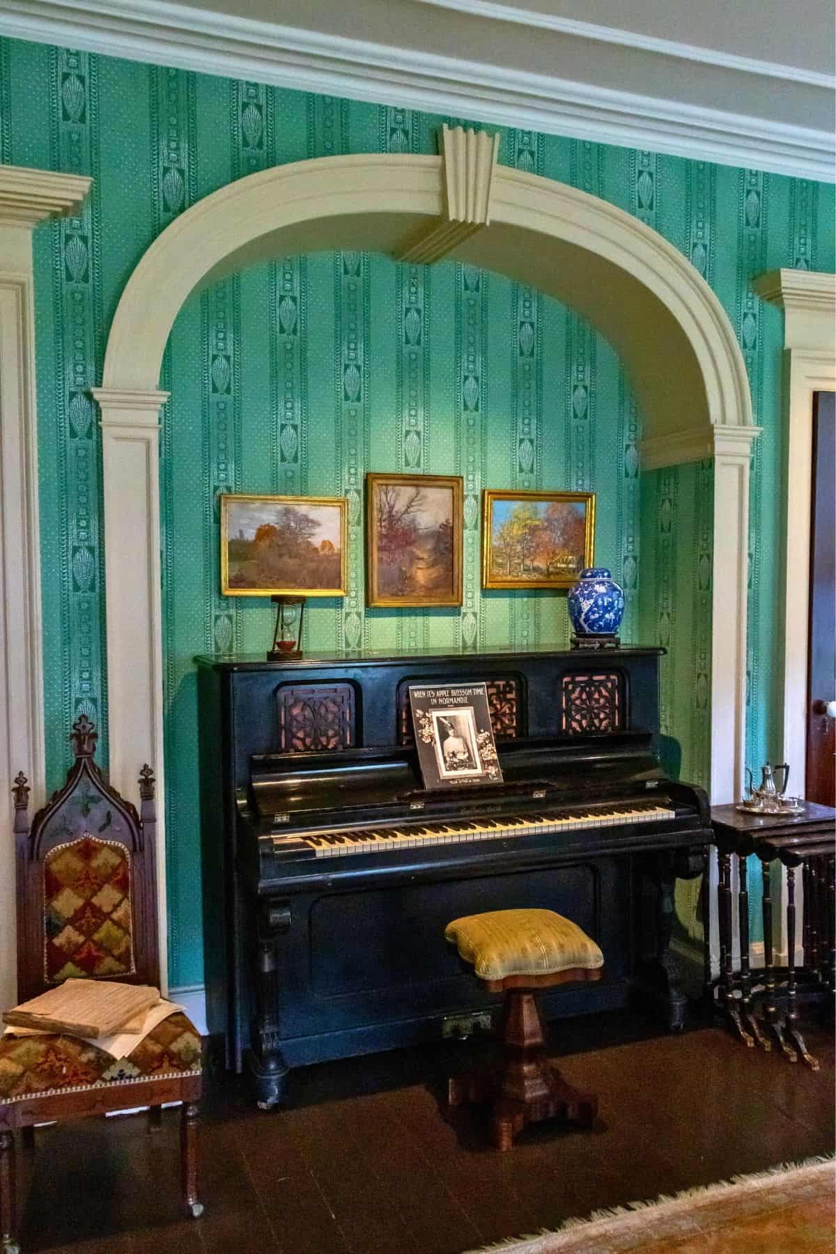 An upright piano stands against a green patterned wall, topped with framed pictures and a vase, flanked by two chairs and set under an arched alcove.