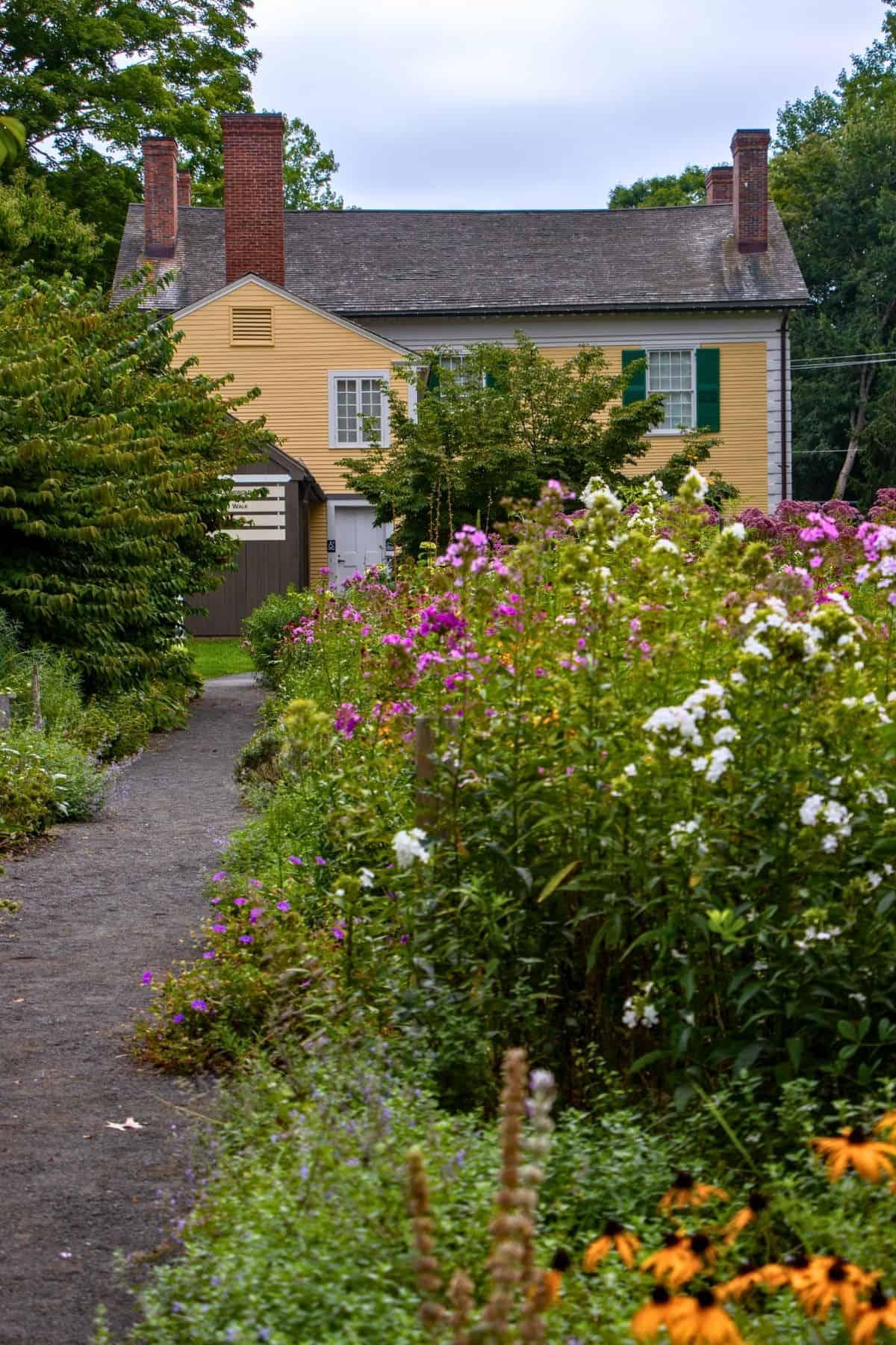 A yellow house with two chimneys is seen behind a garden path lined with colorful wildflowers and green bushes.