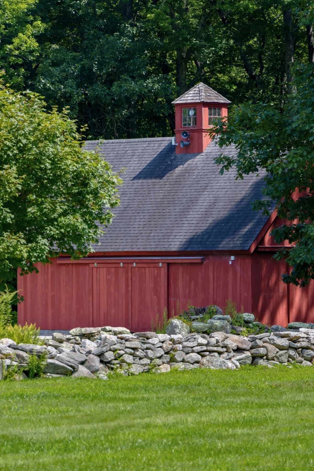 A red wooden barn with a small cupola on the roof is partially obscured by trees, with a stone wall and green grass in the foreground.