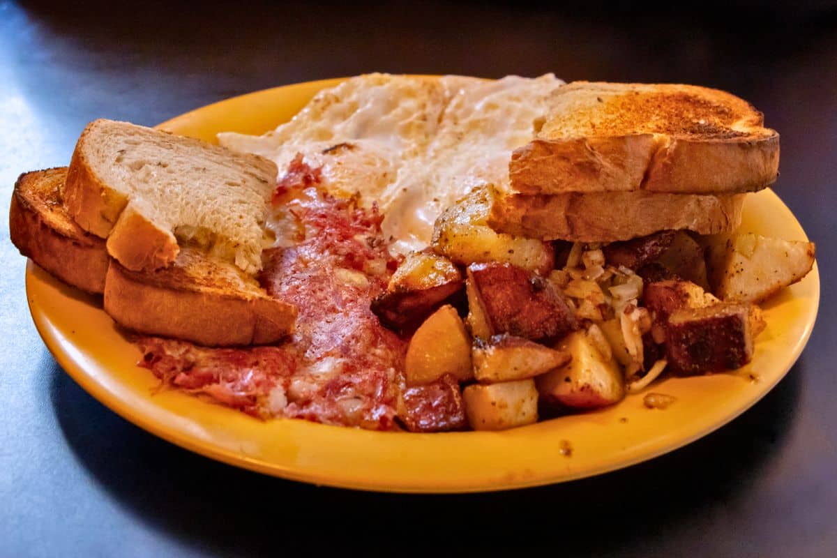 A breakfast plate with two fried eggs, corned beef hash, home fries with onions, and three slices of toasted bread on a yellow plate.