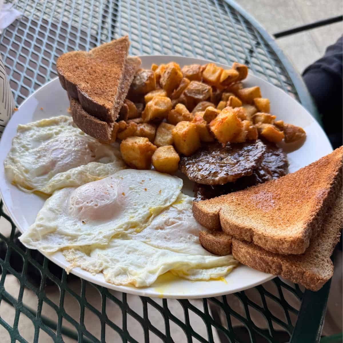 A plate with three fried eggs, toast slices, breakfast potatoes, and two sausage patties sits on a metal outdoor table.