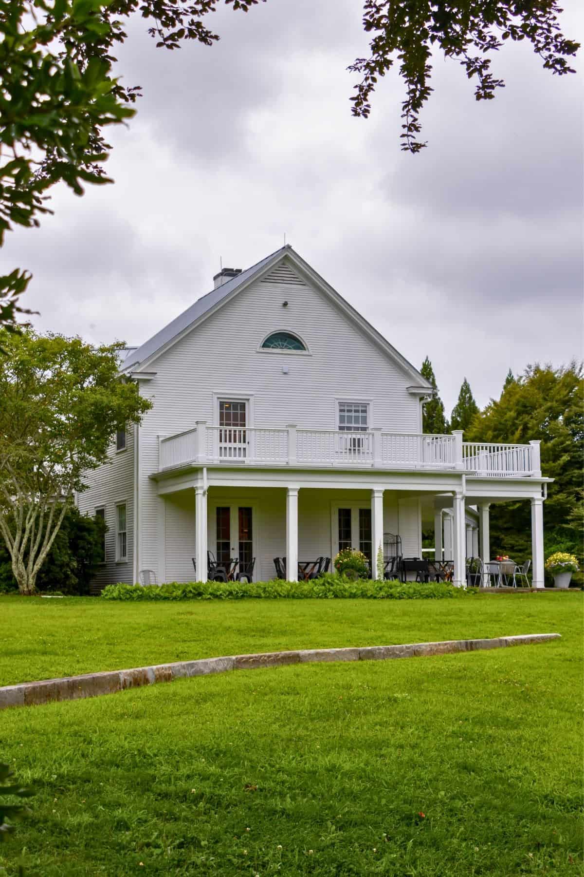 Two-story white house with a large balcony and covered porch, surrounded by green lawn and trees under a cloudy sky.