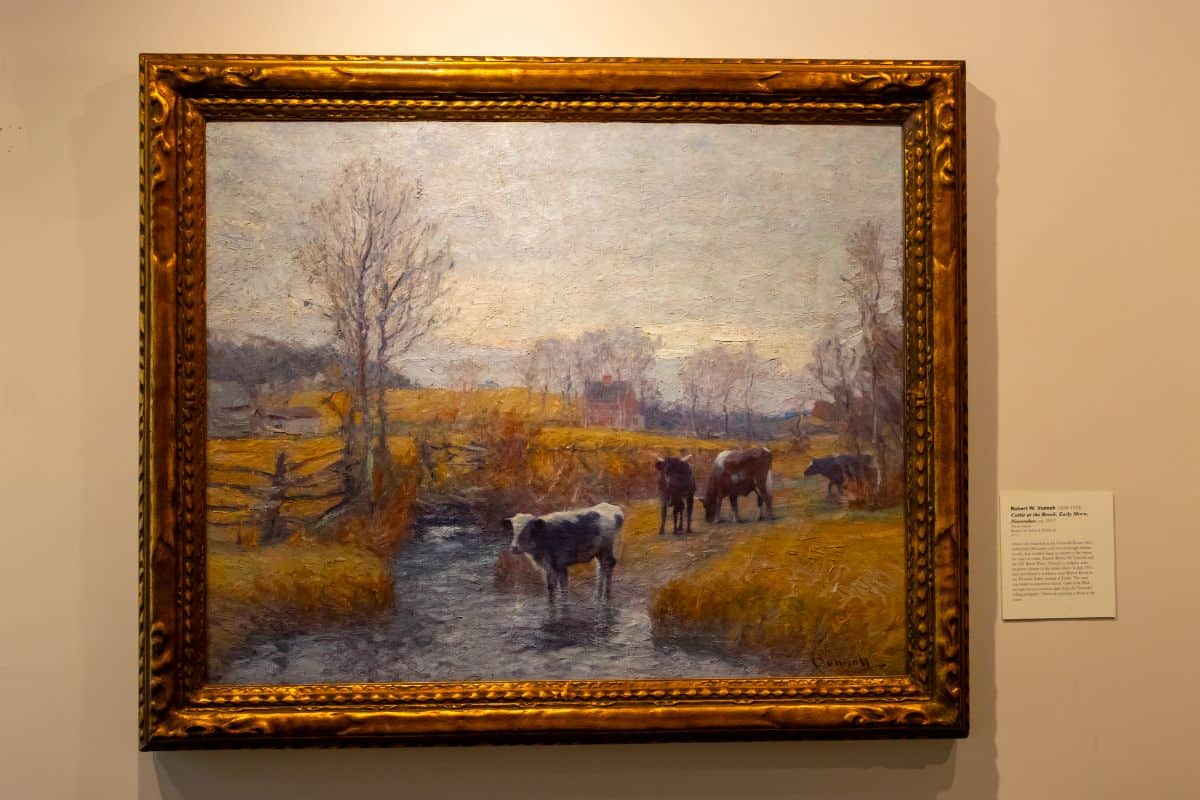 A framed oil painting depicts cows drinking from a stream in a rural landscape with bare trees and a house in the background. A placard with information is mounted on the wall beside the artwork.