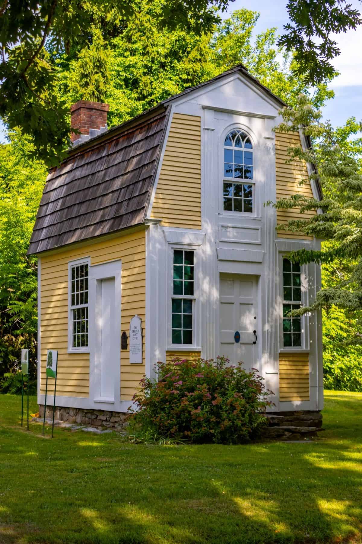 Small yellow house with white trim, shingle roof, and symmetrical windows, surrounded by green trees and grass on a sunny day.